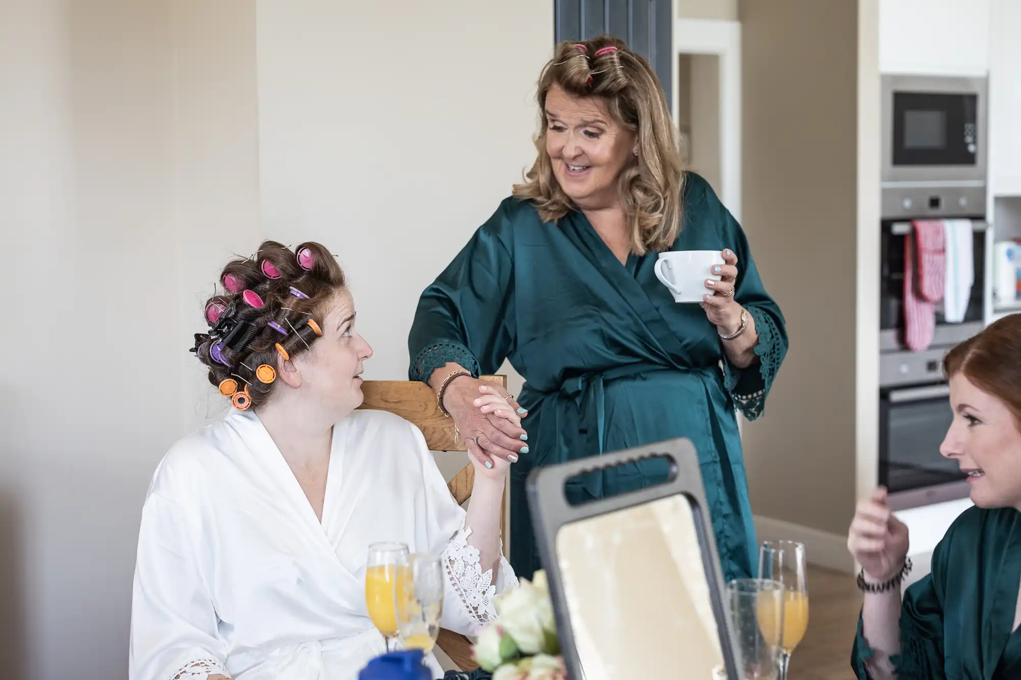 Three women in robes are in a home kitchen. One woman with hair rollers holds hands with a woman holding a coffee cup as they smile at each other. A third woman sits nearby with a glass of orange juice.