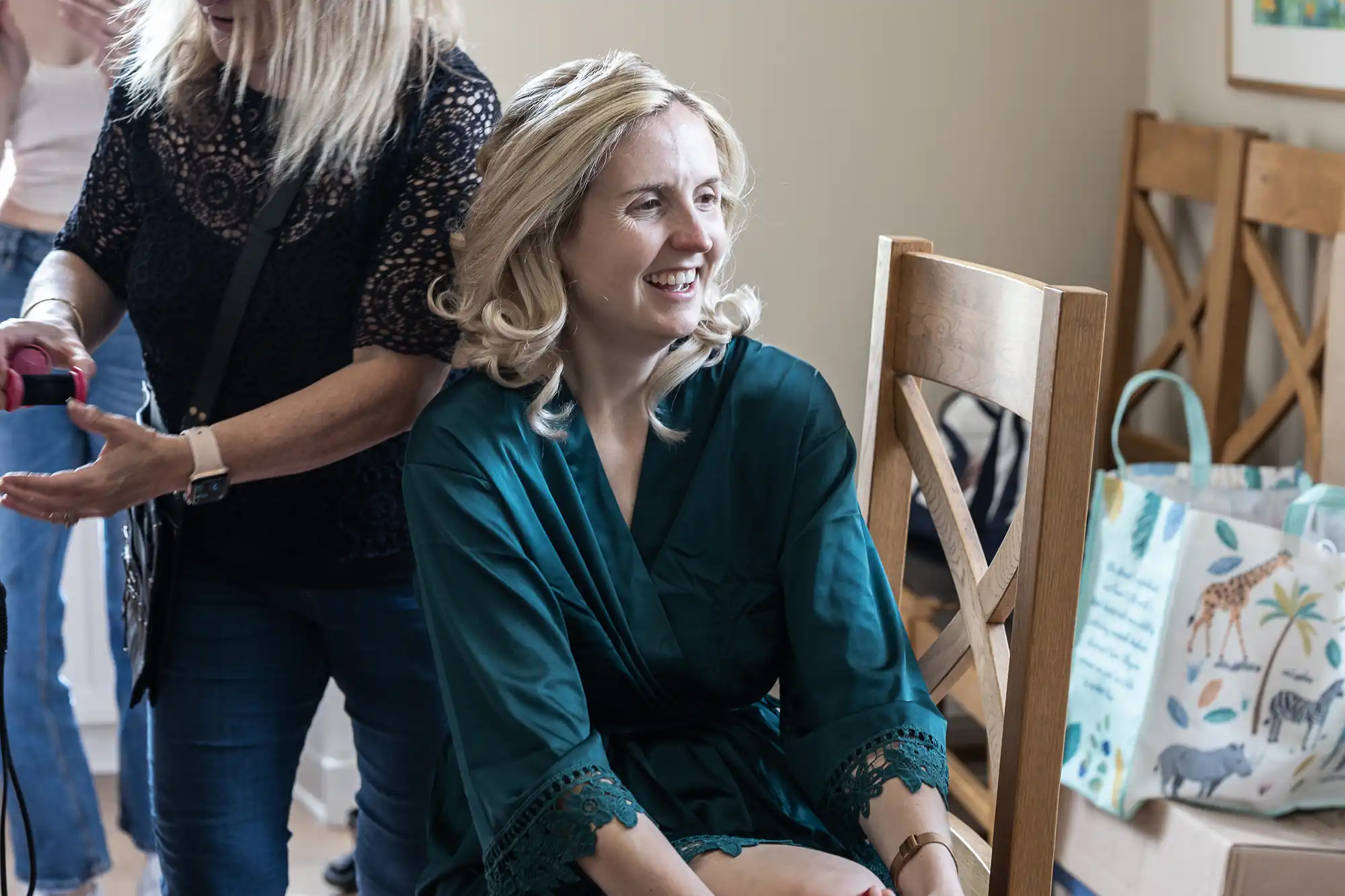 A woman with blonde hair, wearing a green robe, sits on a wooden chair and smiles. Another woman is standing beside her, holding a curling iron.