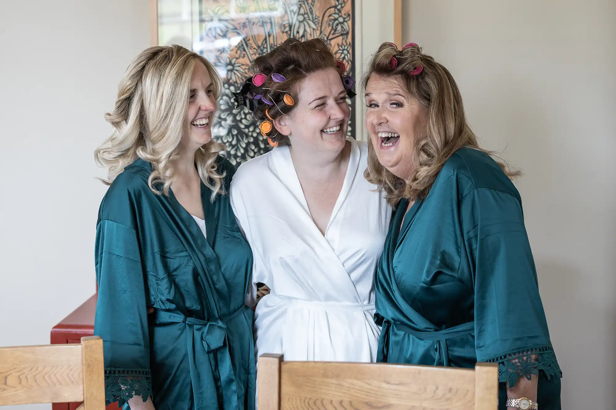 Three women in robes, two in teal and one in white, laugh together indoors, with hair curlers in their hair.