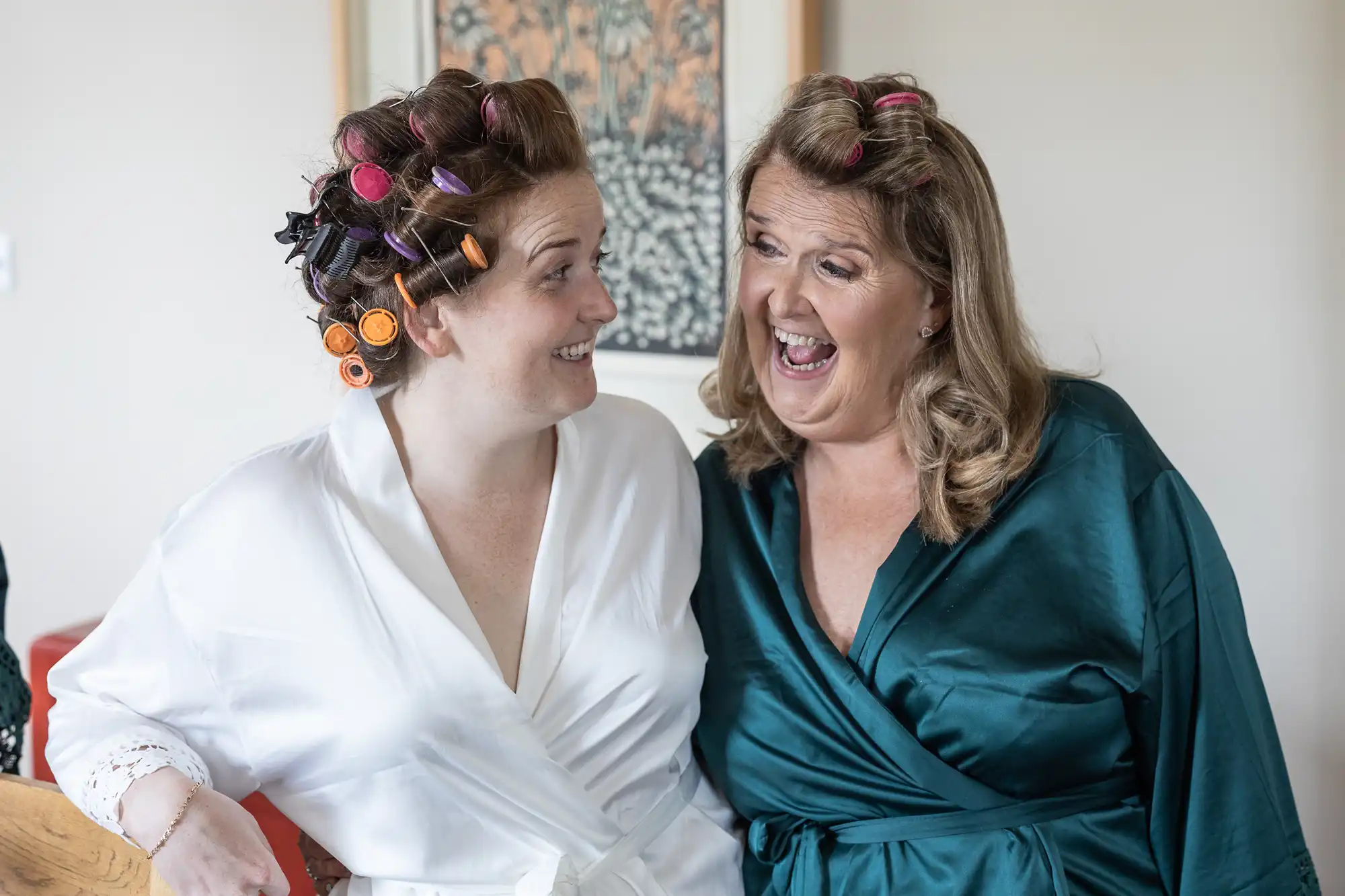 Two women in robes smile and laugh with hair rollers in their hair. They appear to be getting ready together in a relaxed, joyful atmosphere.