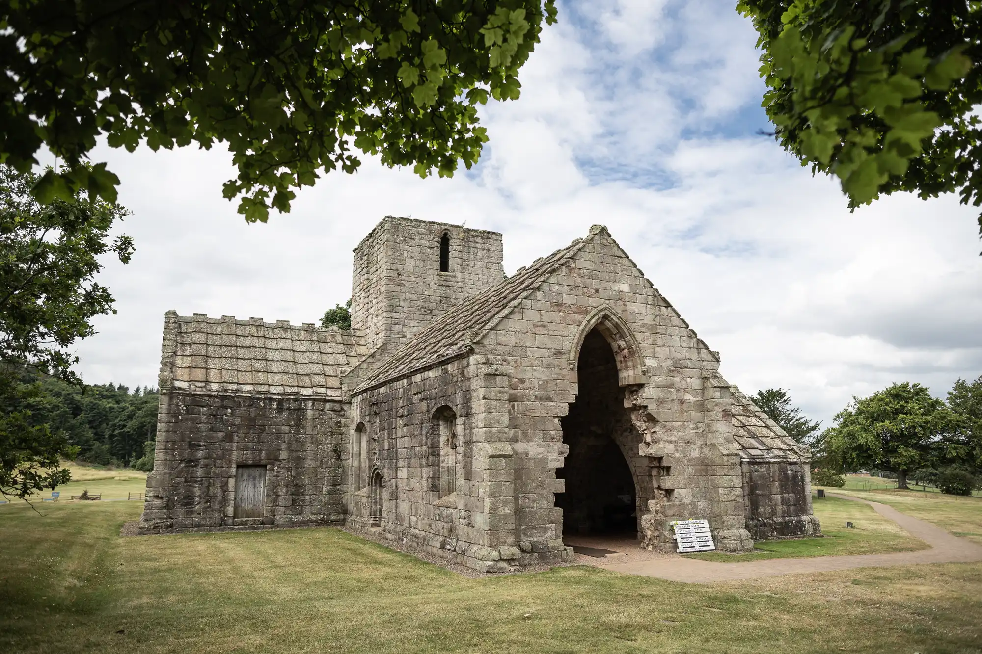 A stone church with a rustic architecture featuring an arched entrance, surrounded by green trees and a grassy lawn under a cloudy sky.