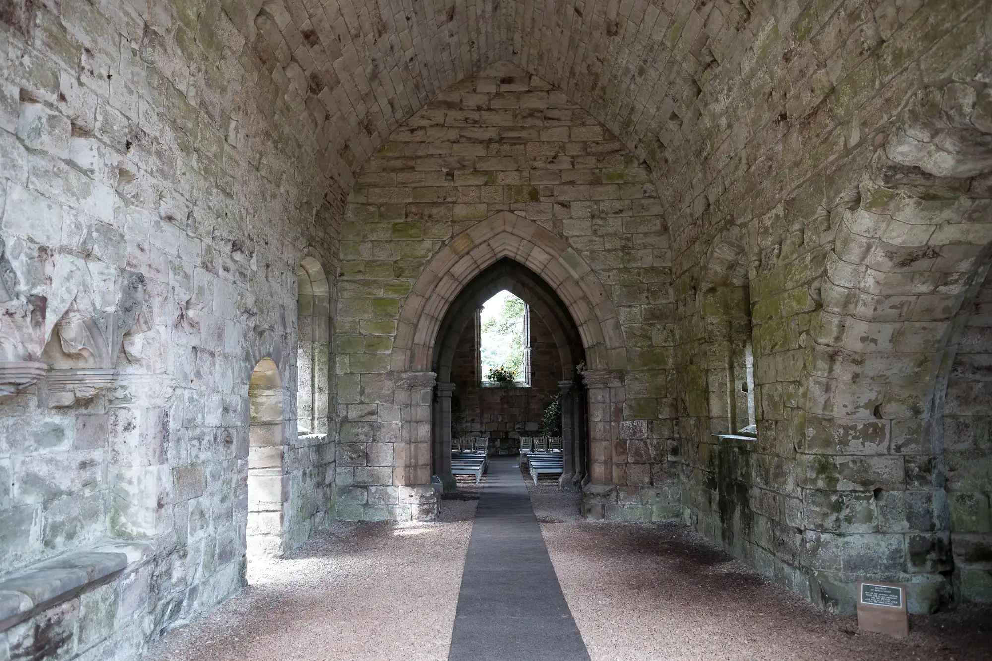 A stone archway inside a historic building with moss-covered walls and a small window at the far end, allowing natural light to filter through. The floor is covered in gravel and a pathway.