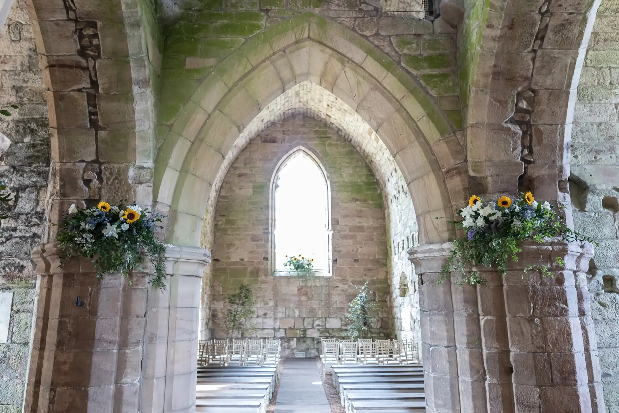 Stone archways and pews in an old chapel with flower arrangements on the columns. A bright window is at the back, allowing light to enter the room.