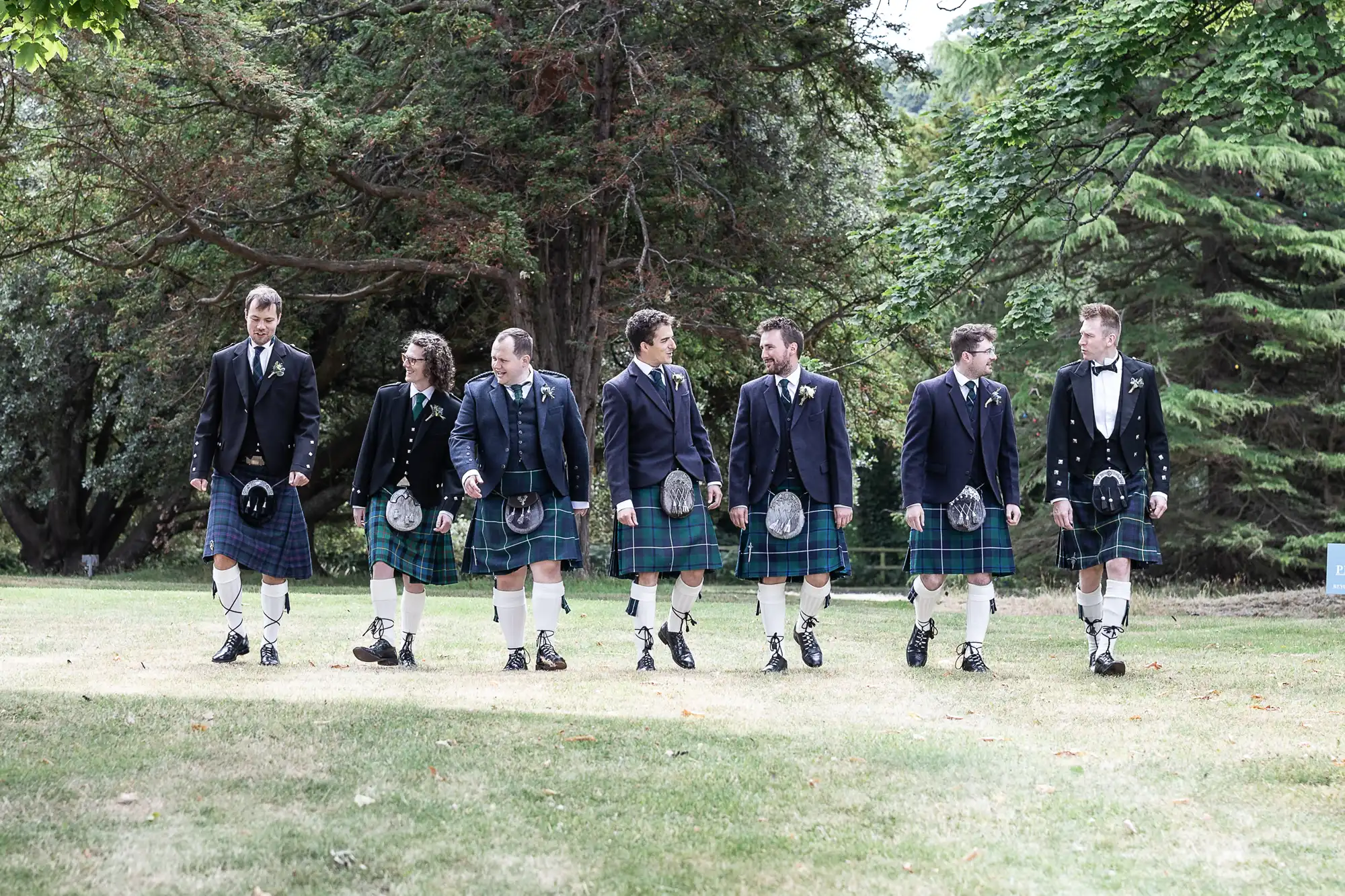 A group of seven men wearing kilts and formal jackets walk on grass in an outdoor setting with trees in the background.