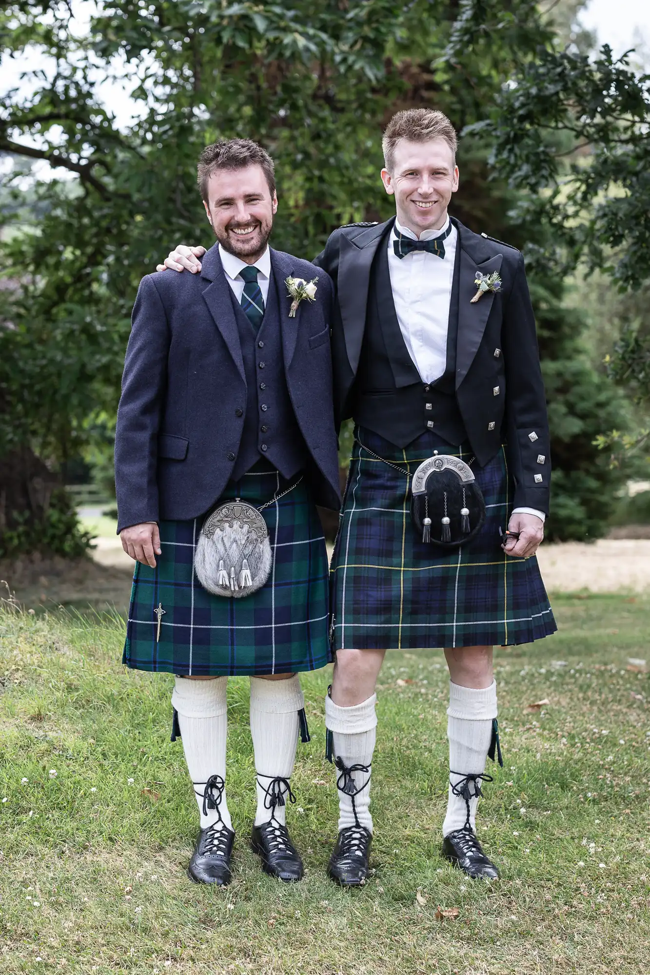 Two men wearing traditional Scottish kilts, jackets, and socks pose together outdoors on a grassy area with trees in the background.