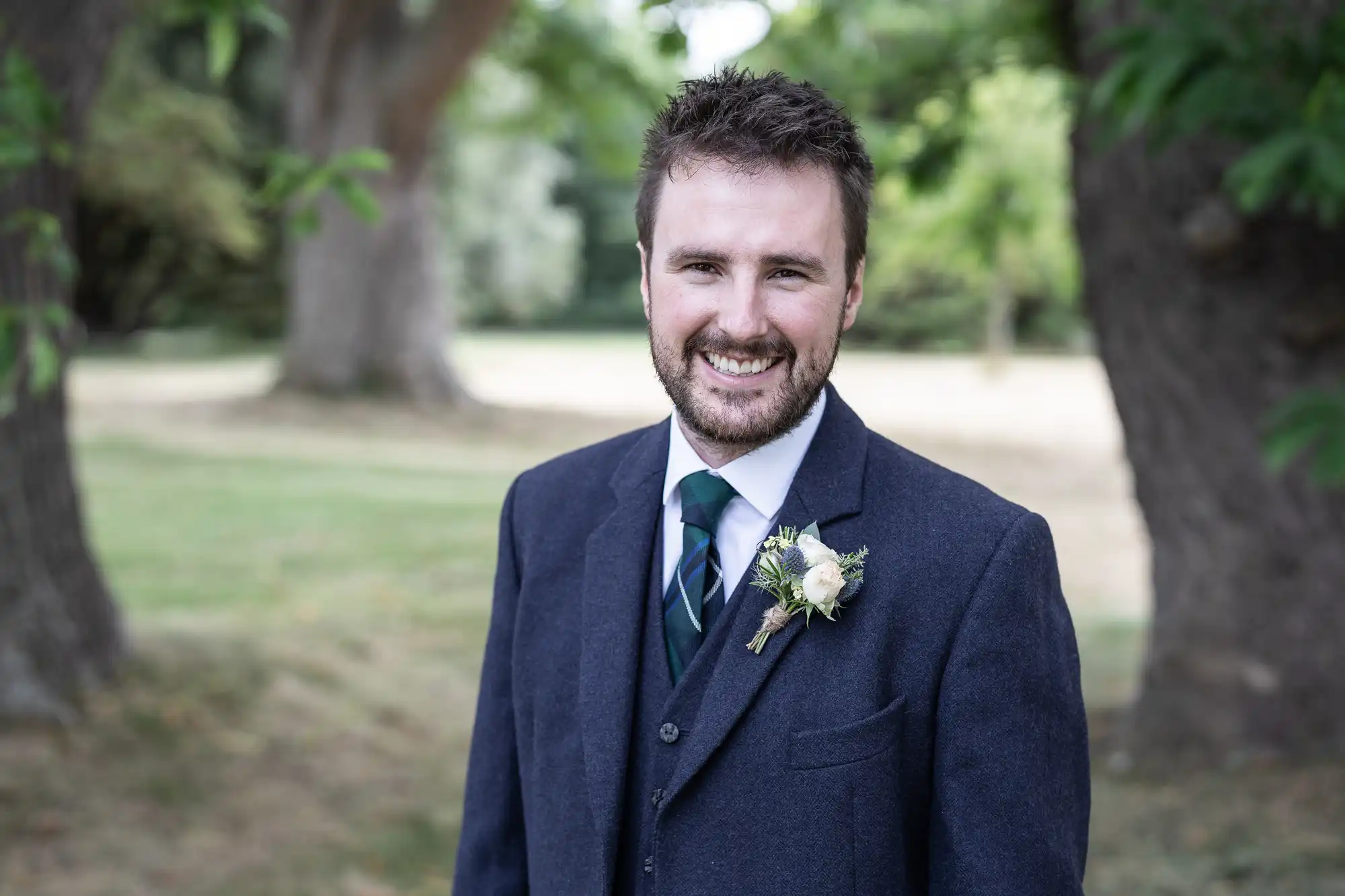 A man in a dark suit with a boutonniere smiles while standing outdoors in a park with trees in the background.