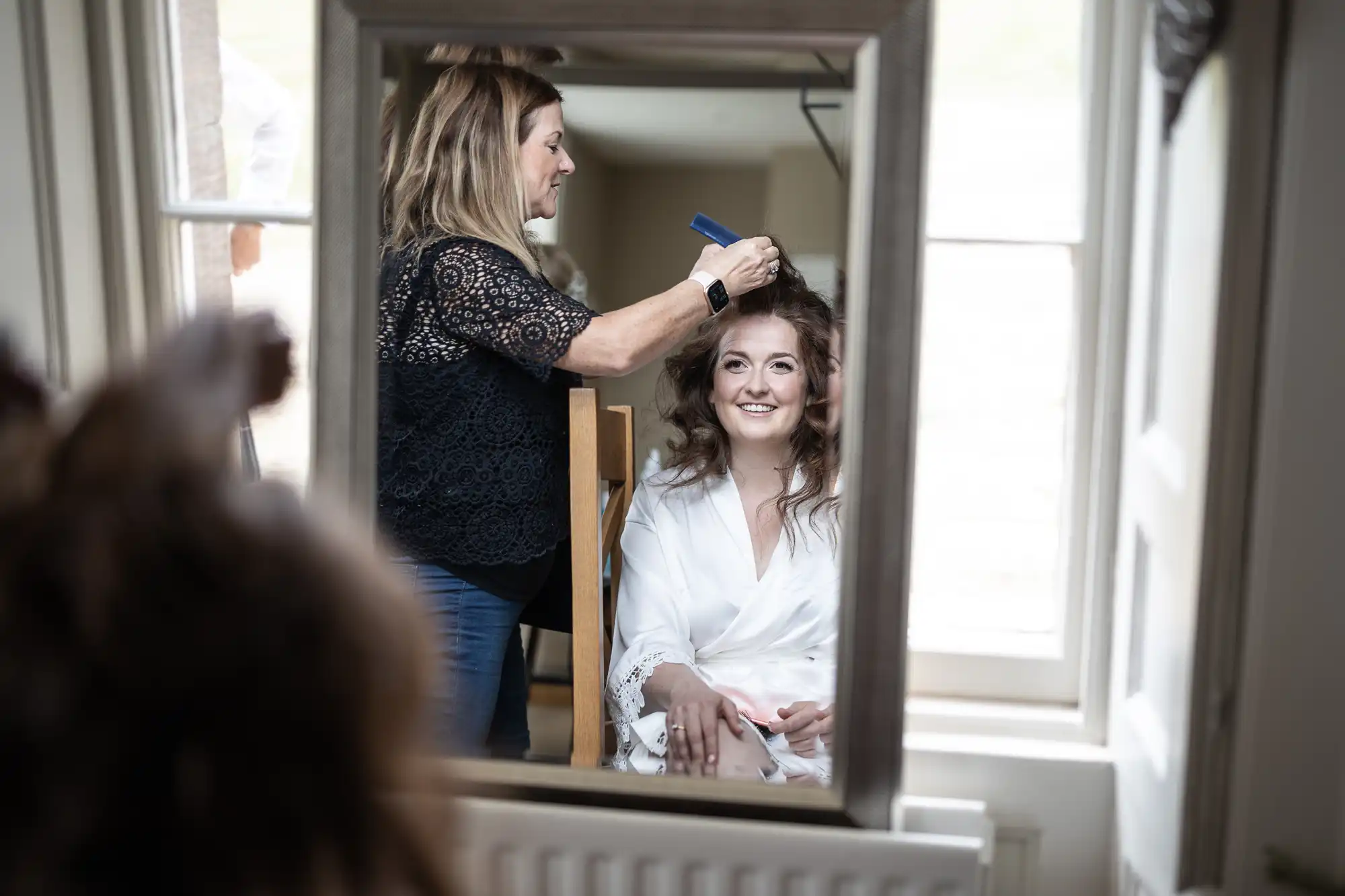 A woman gets her hair styled by a hairdresser, as seen in a mirror reflection. She is smiling and seated wearing a white robe.