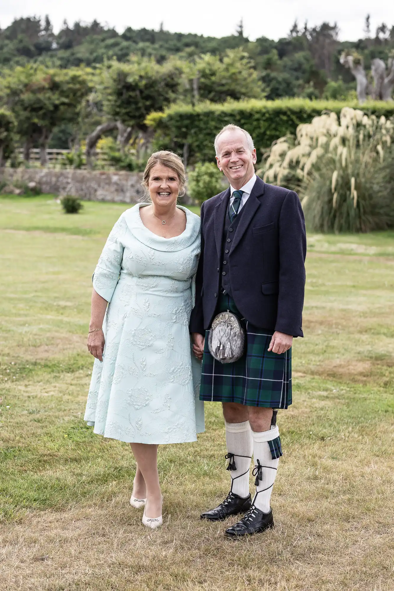 A couple stands together smiling outdoors, the woman in a light blue dress and the man in a traditional Scottish outfit with a kilt. Trees and a lawn are in the background.