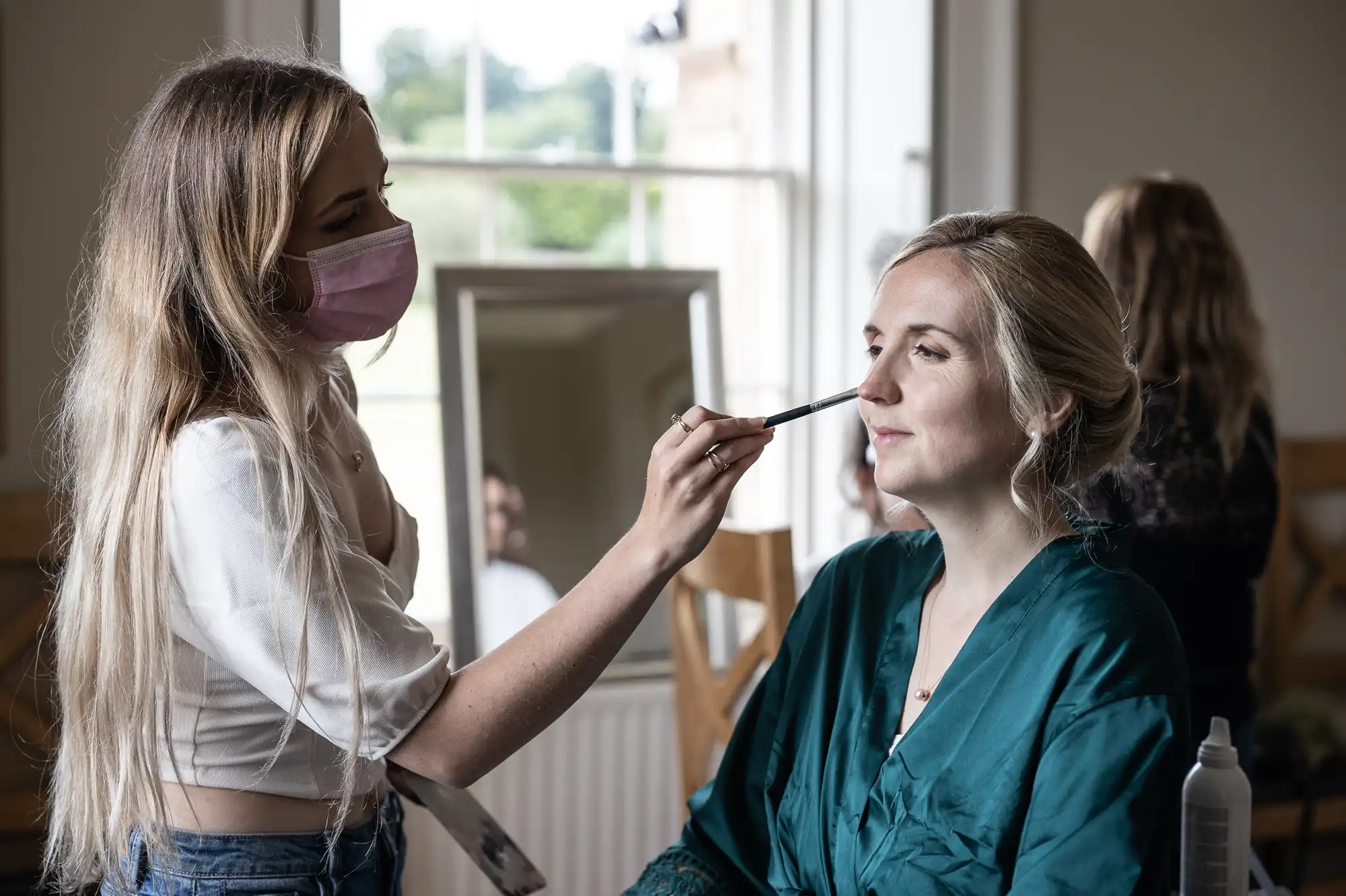 A woman in a green robe gets her makeup done by a woman wearing a pink face mask. They are in a well-lit room with a window and mirror in the background.