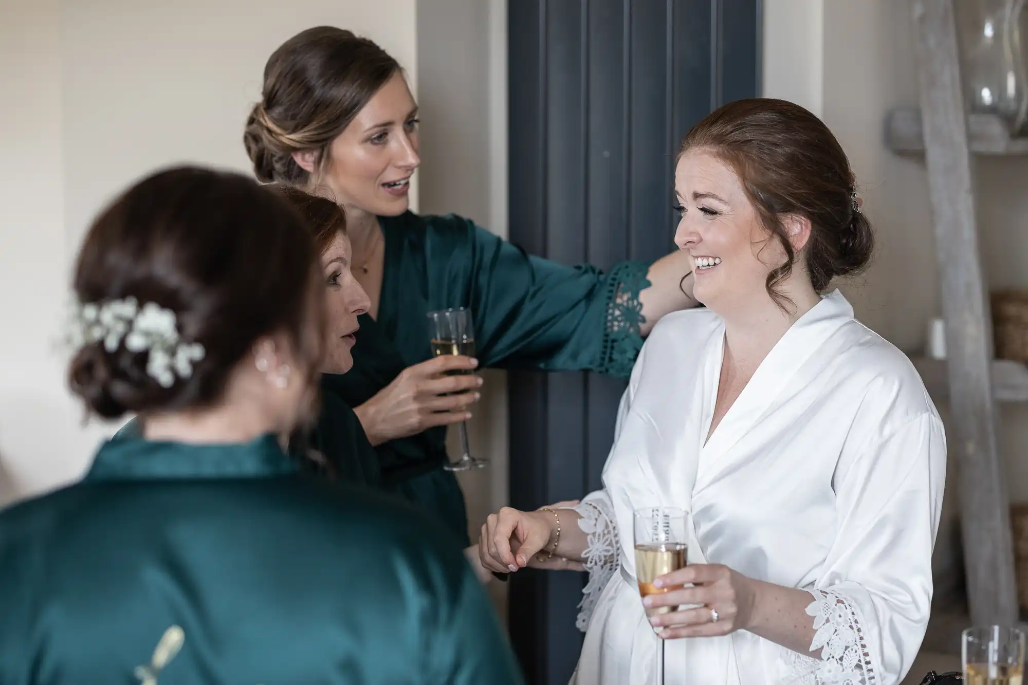 A group of women in robes, drink champagne and converse cheerfully. One woman in a white robe appears to be the center of attention.