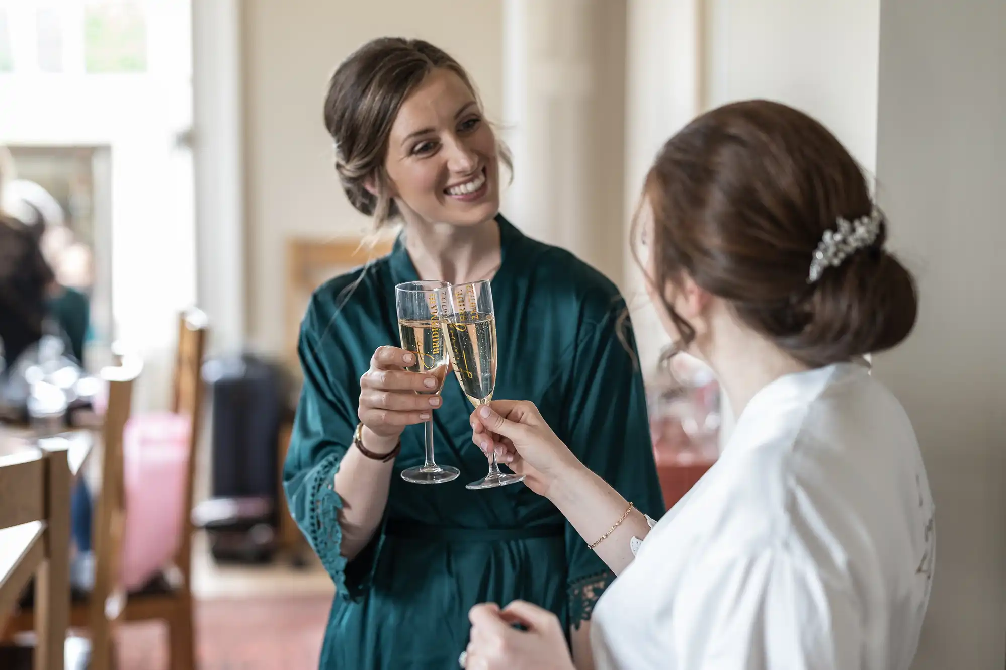 Two women in robes clink champagne glasses in a brightly lit room, smiling at each other.