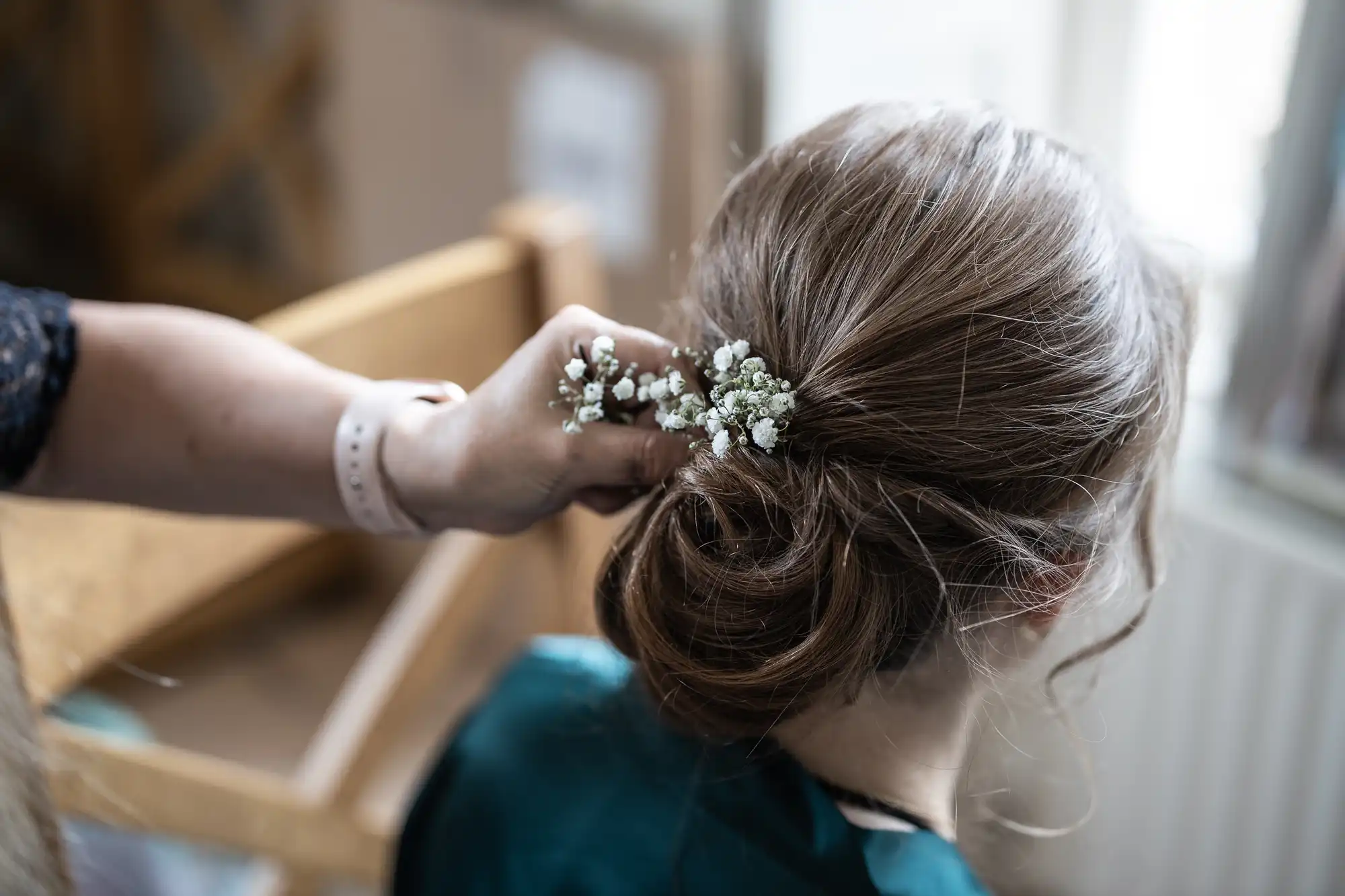 A person is arranging delicate white flowers into a woman's loose, low bun hairstyle.