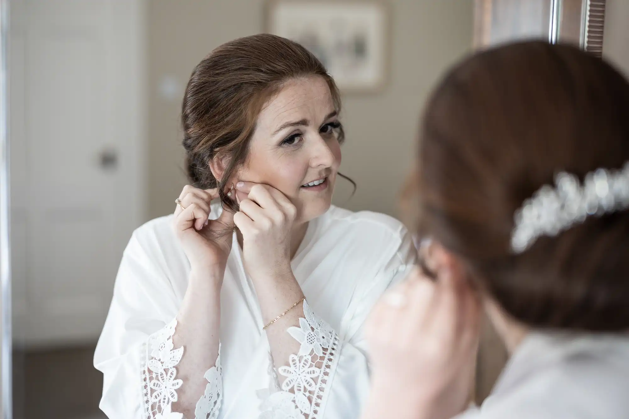 A woman in a white robe with lace sleeves adjusts her earring while looking into a mirror. Her hair is neatly styled in an updo.