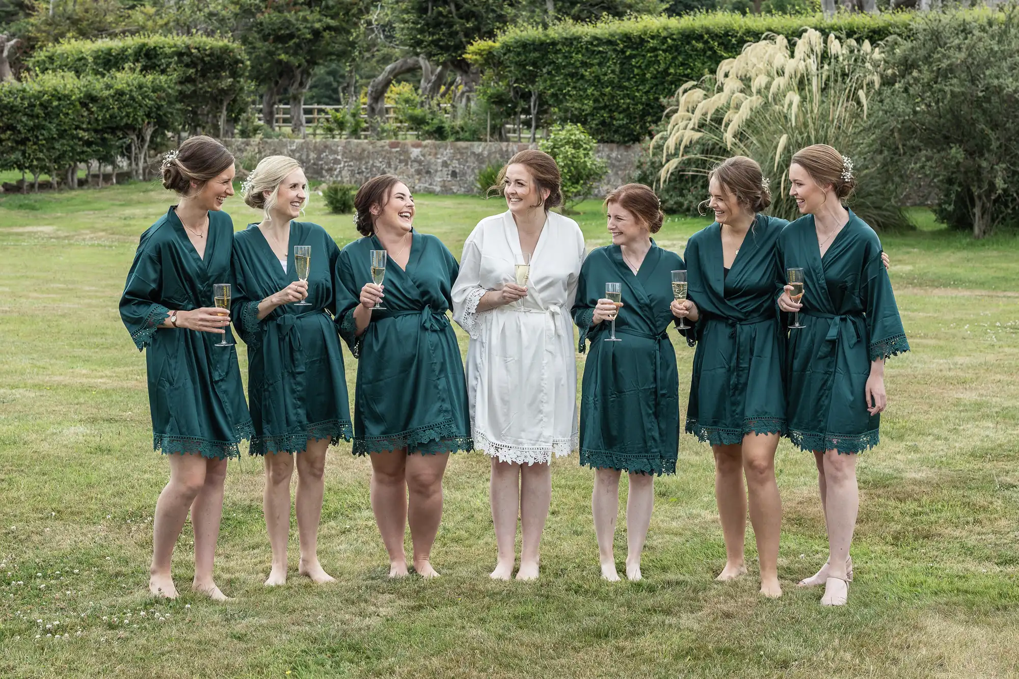 Seven women, six in matching green robes and one in a white robe, stand barefoot on grass, smiling and holding champagne glasses.