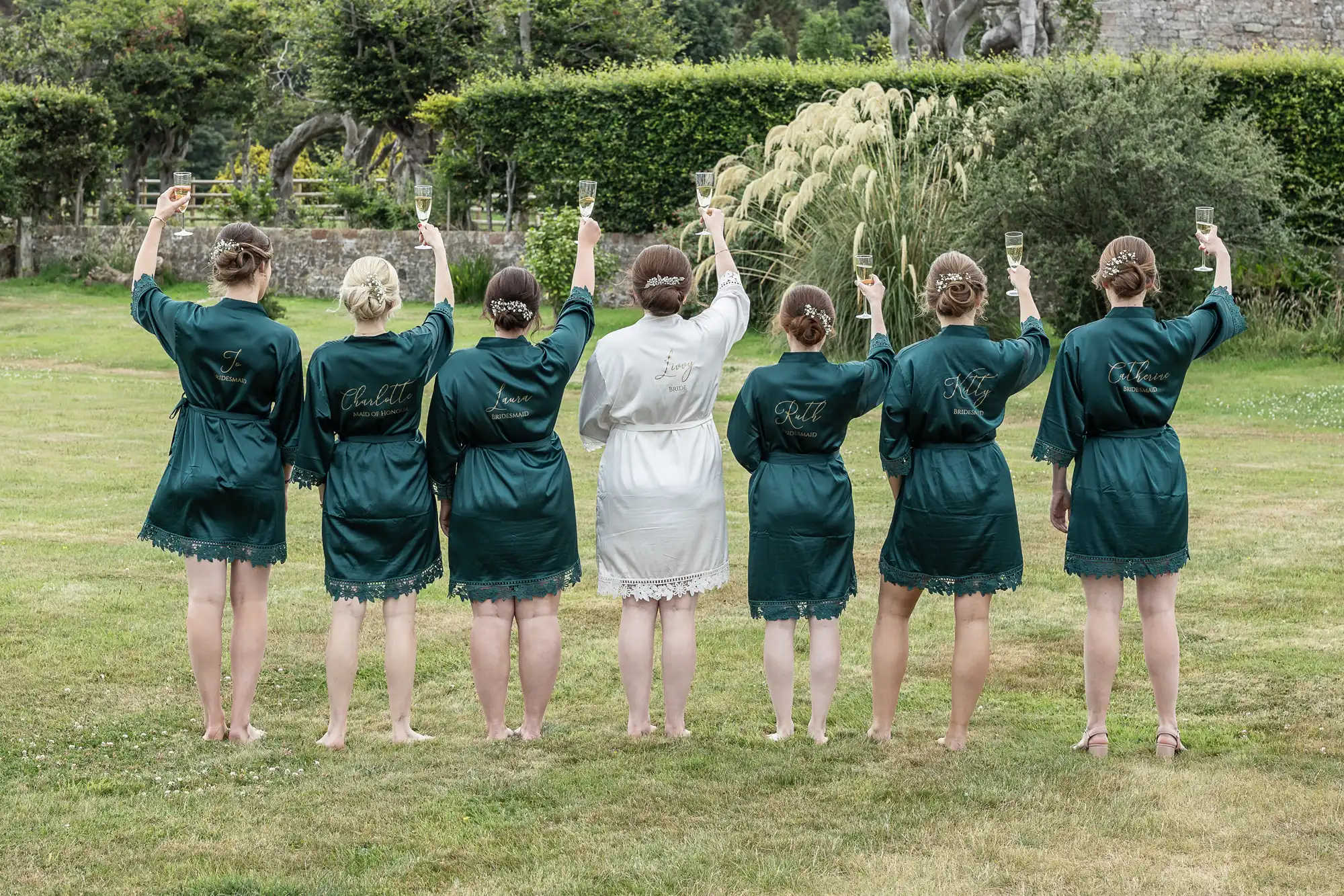 Seven people in robes stand with their backs to the camera, raising glasses of champagne in a garden area.