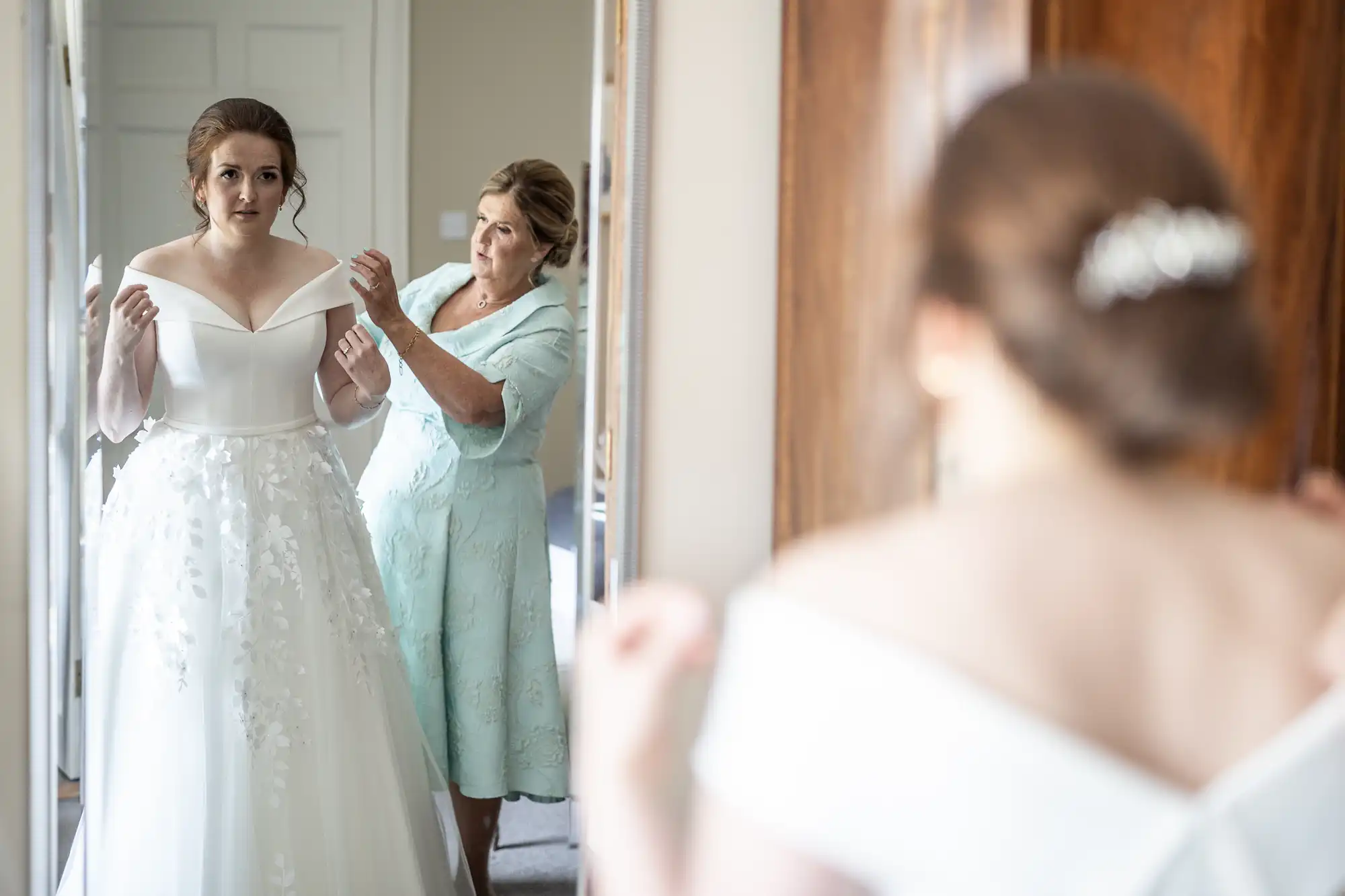 A bride in a white dress is adjusting her sleeves while an older woman helps her. They are standing in front of a mirror.