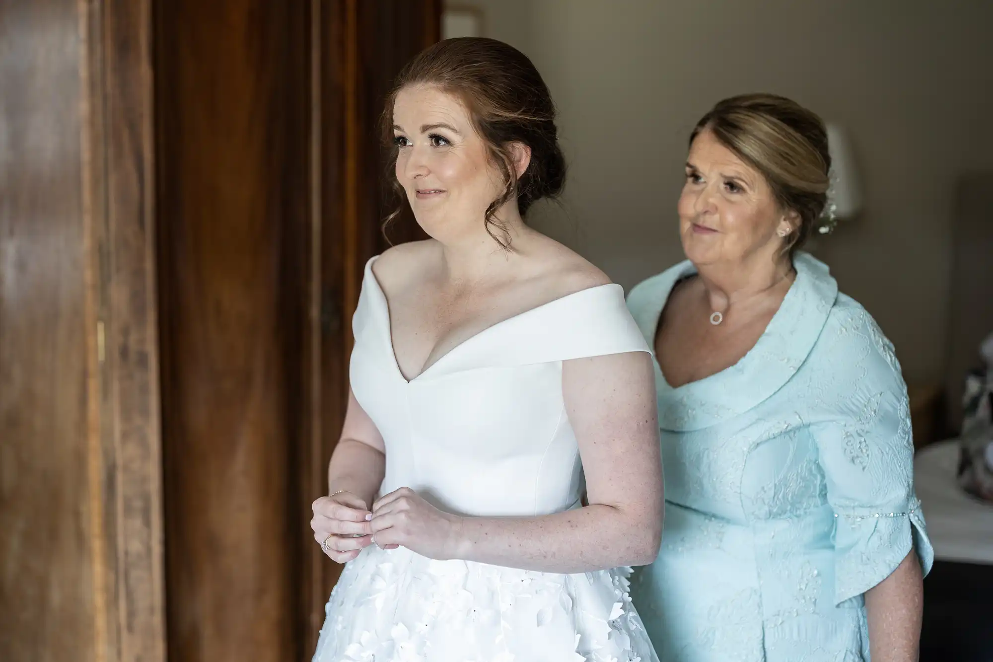 A bride in a white dress stands inside a room with a woman in a light blue outfit standing behind her, both looking towards the left.