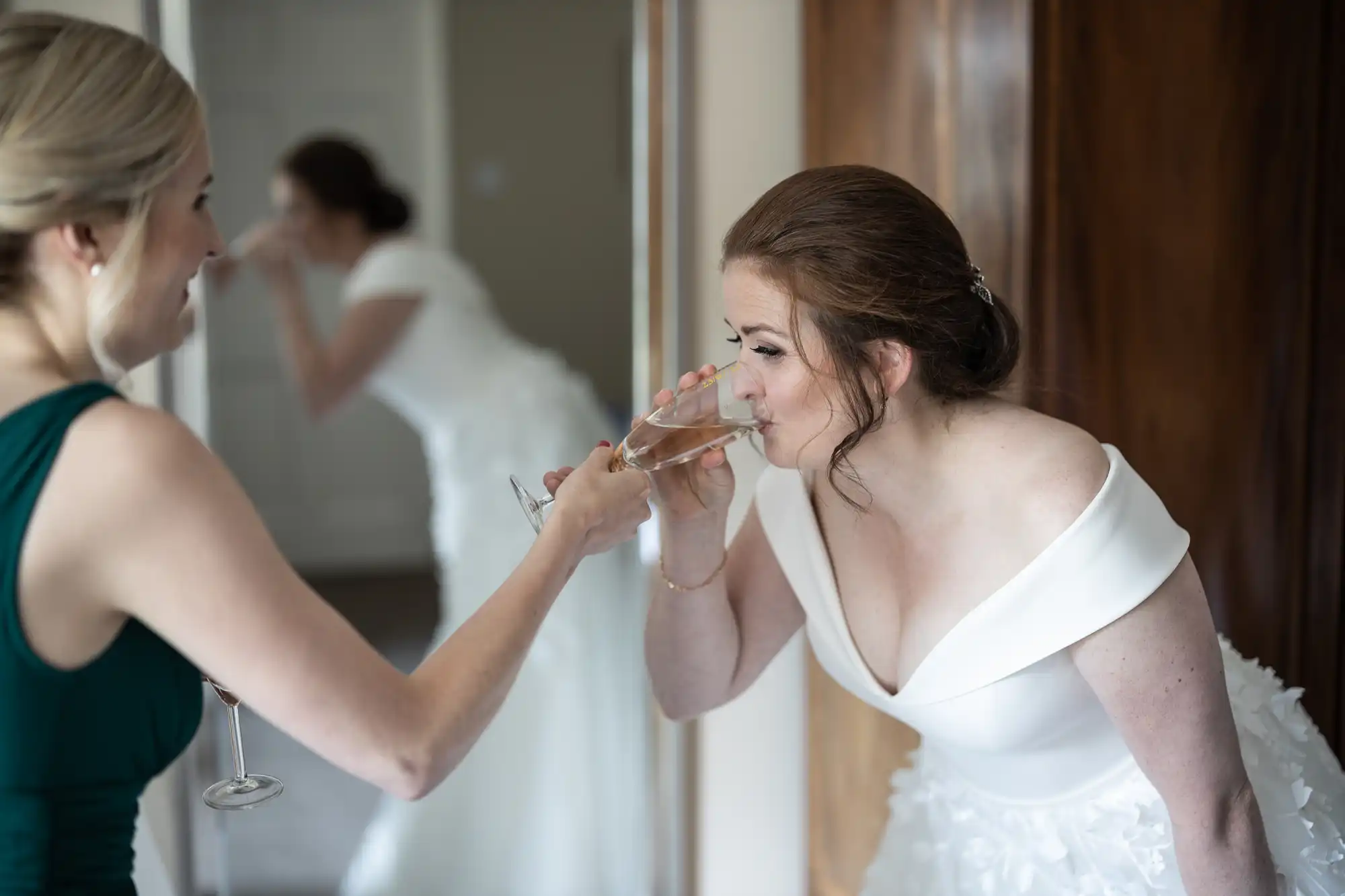 A bride in a white dress drinks from a champagne flute held by a woman in a green dress in a room with a mirror.