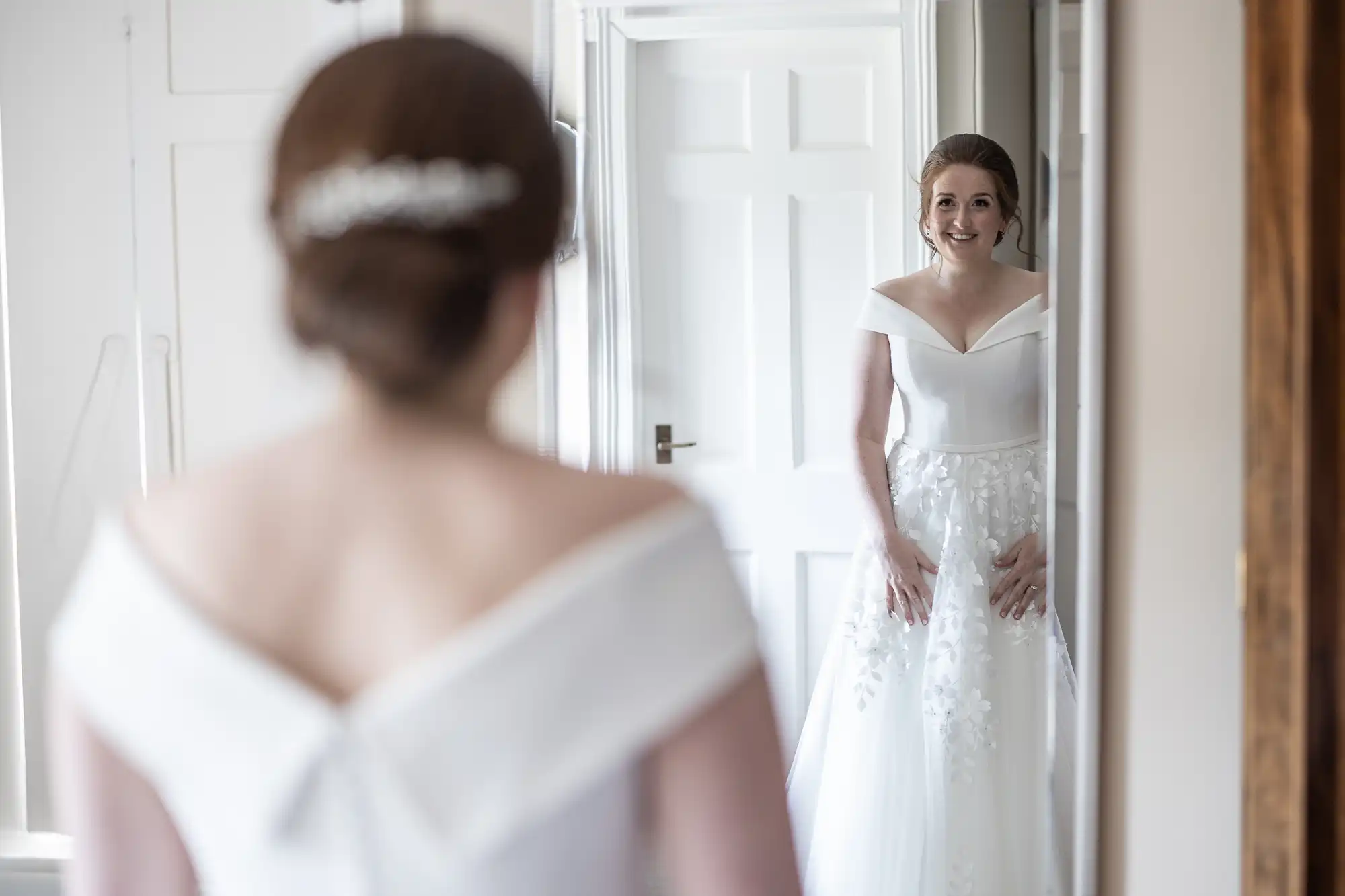 A bride in an off-the-shoulder white wedding dress stands in front of a mirror, smiling at her reflection.