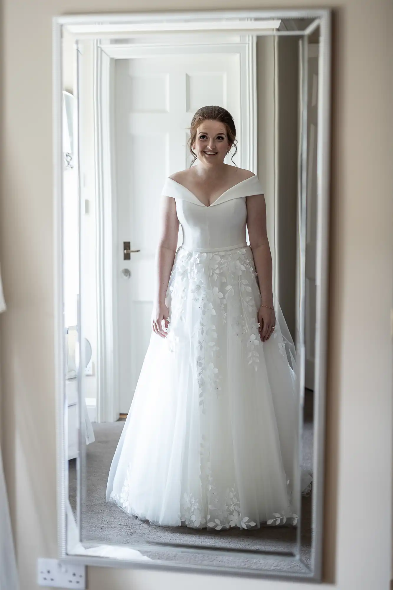 A woman in an off-the-shoulder white wedding dress stands in front of a mirror, smiling.