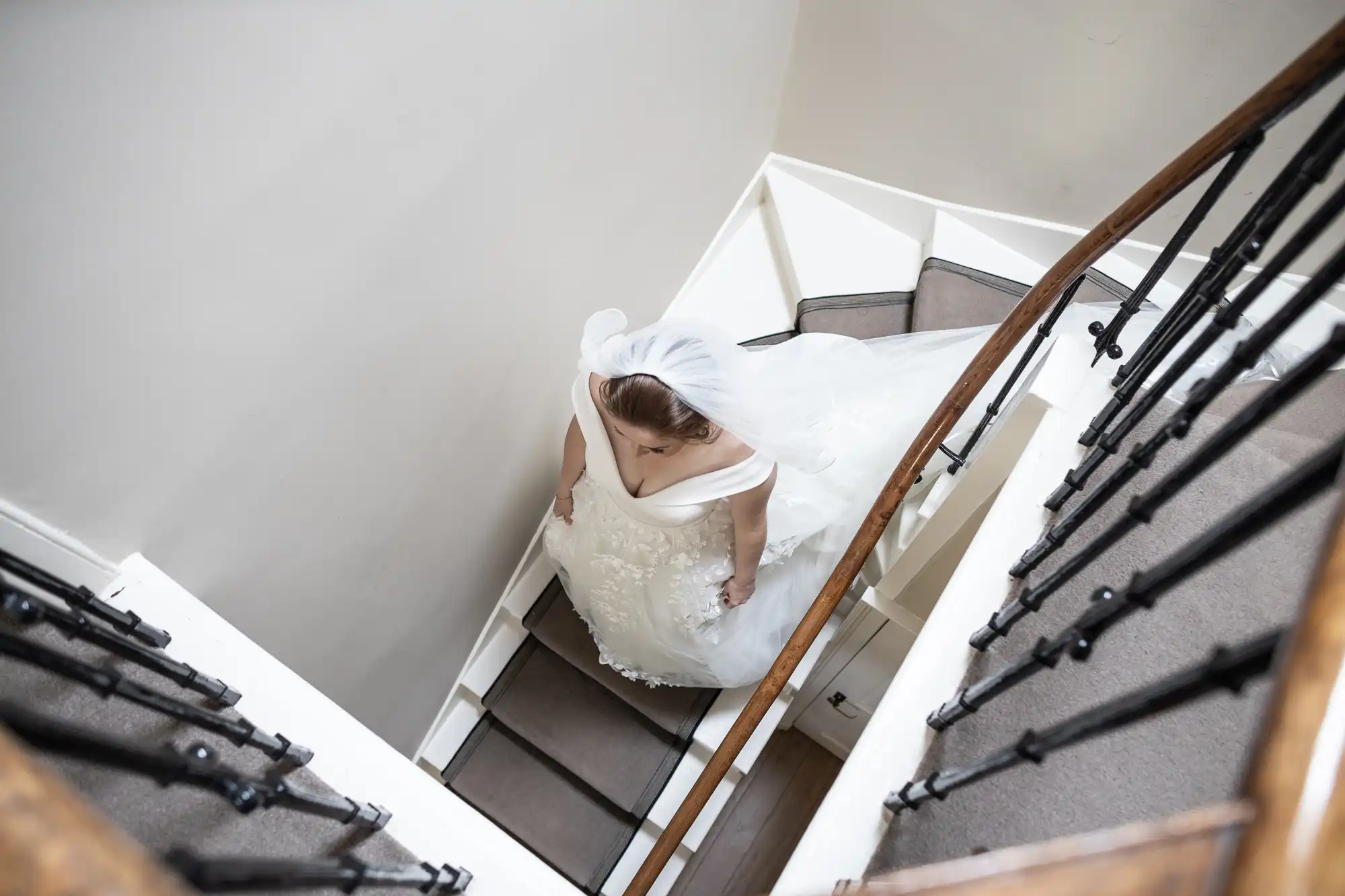 A bride in a white wedding dress and veil descends a narrow, winding staircase with black railings and beige steps.