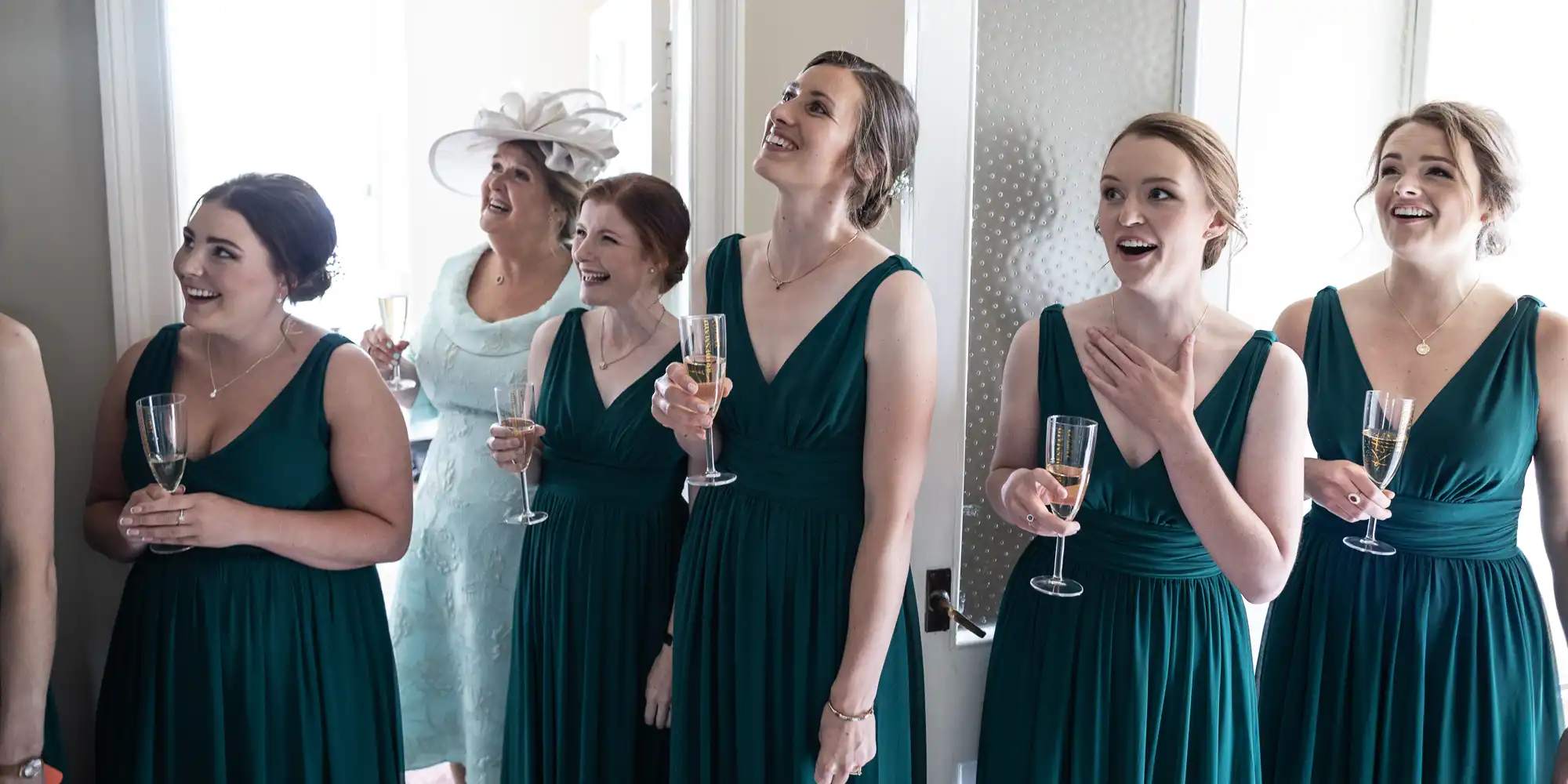 A group of six women, five in dark green dresses and one in light blue, holding glasses of champagne, looking surprised and smiling in a bright room.