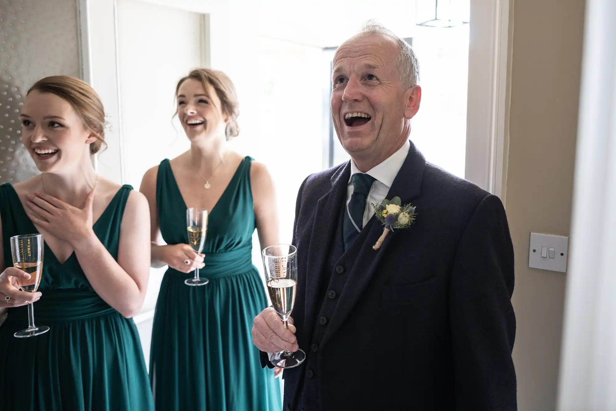 Three people in formal attire, including two women in green dresses holding champagne flutes and an older man in a suit, react with smiles and laughter indoors.