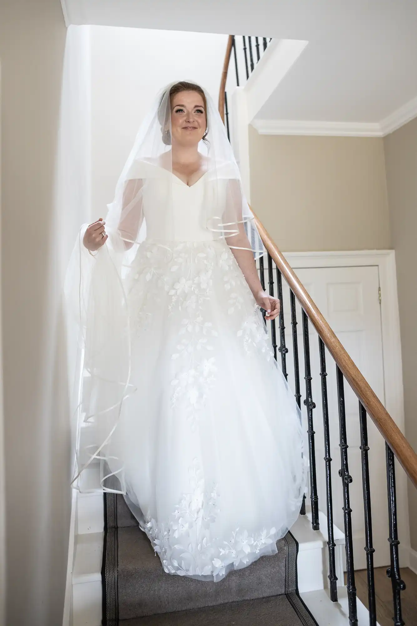 A bride in a white wedding dress and veil is descending a staircase with a wooden handrail inside a house.