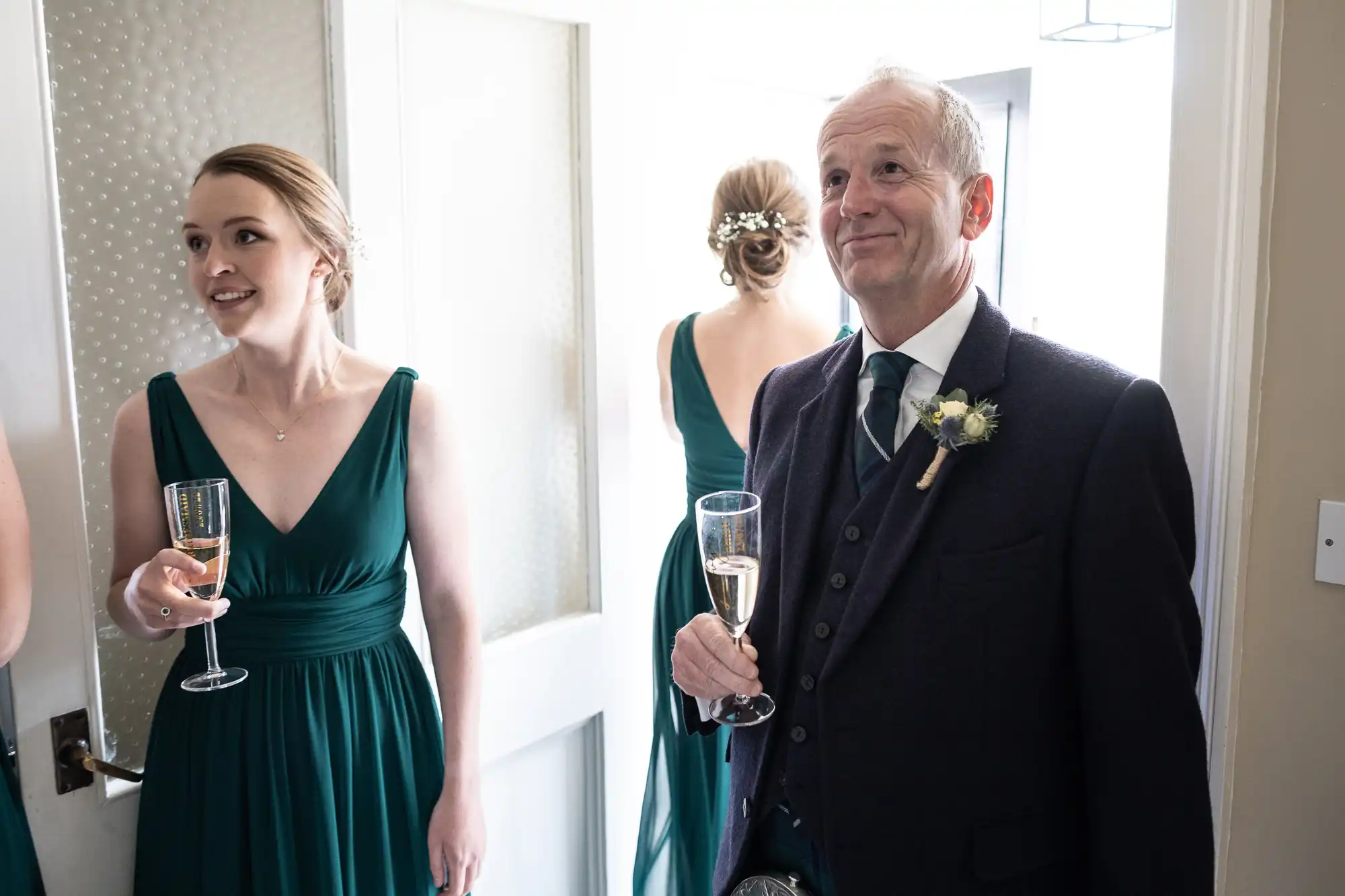 A woman and an older man, both dressed formally, hold champagne glasses and smile. Another woman, her back to the camera, stands nearby.