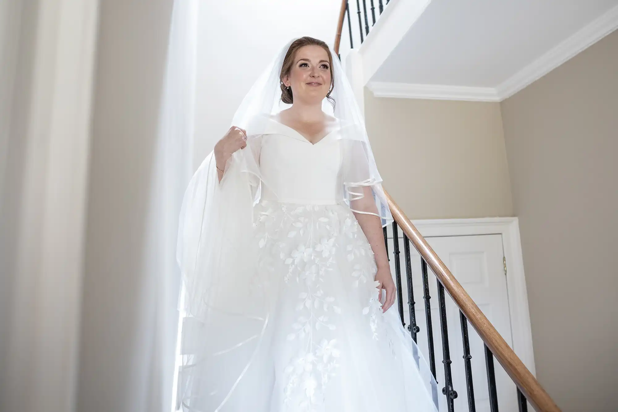 A bride in a white wedding dress and veil descends a staircase indoors.