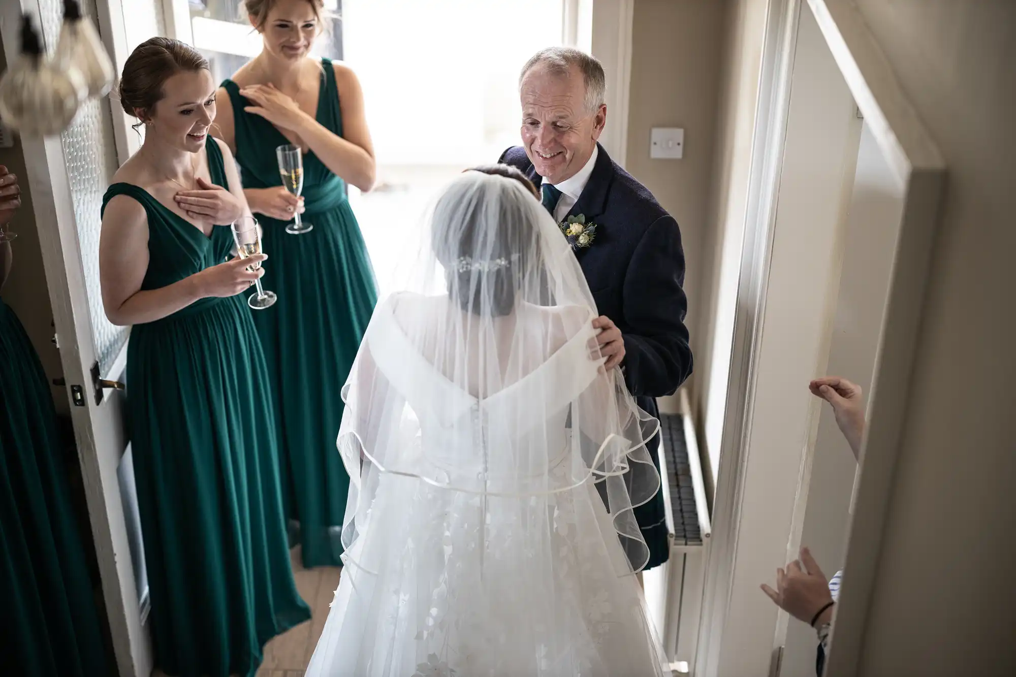 A bride in a white gown and veil is embraced by an older man in a suit, surrounded by three bridesmaids in green dresses, holding glasses of champagne.