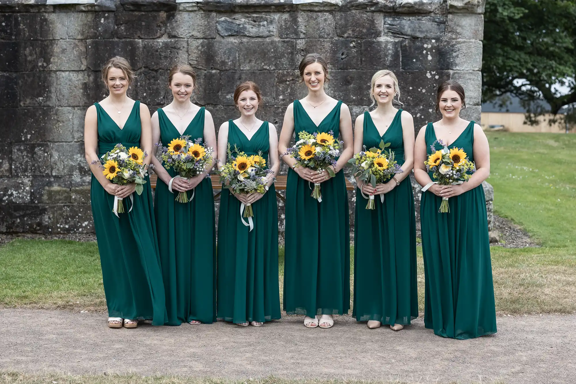 Six women in green dresses stand in a line outdoors, holding bouquets of sunflowers and greenery, with a stone wall in the background.