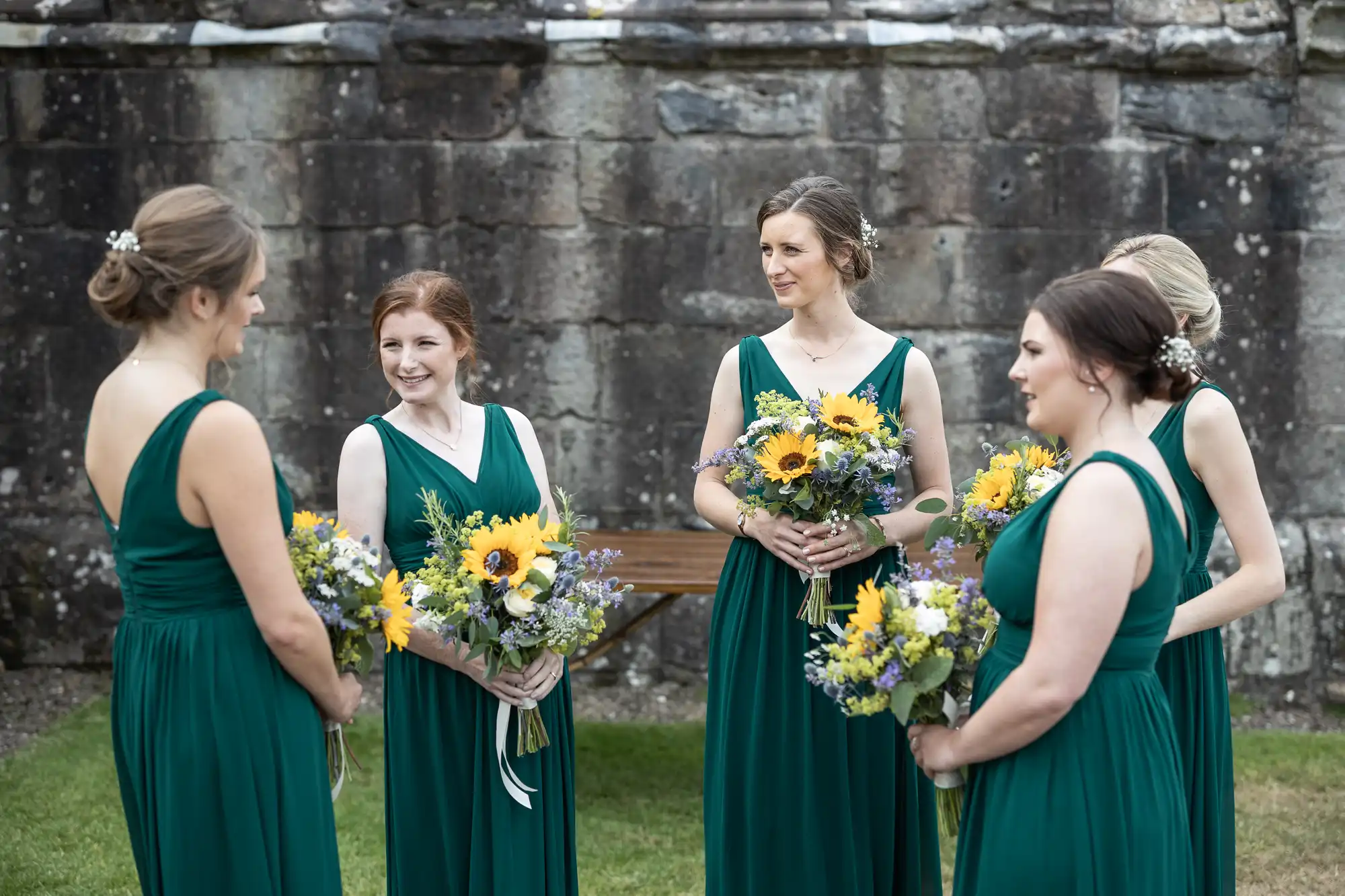 Five women in green dresses stand in a circle outdoors, holding bouquets of sunflowers and other flowers, with a stone wall in the background.