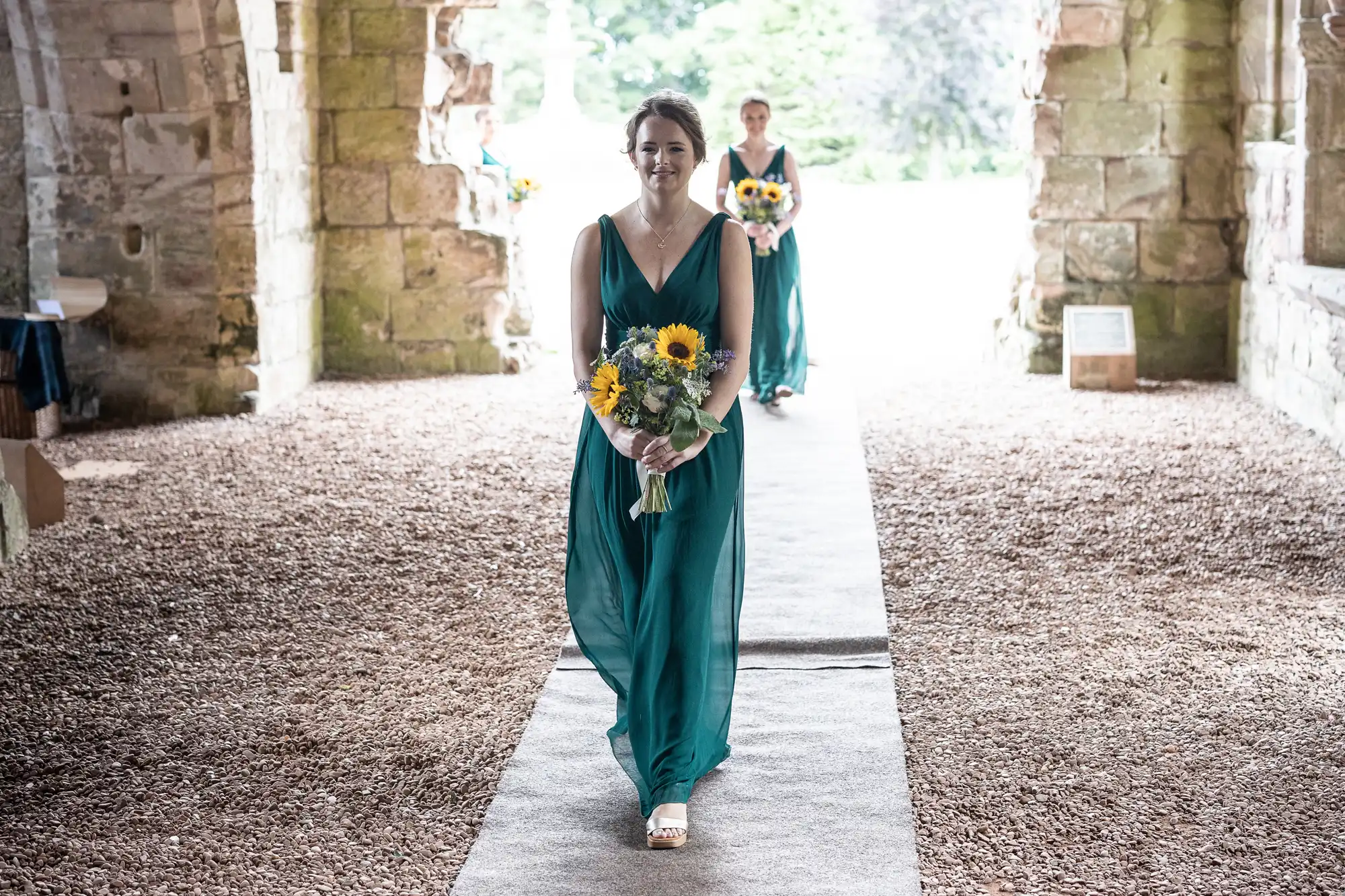 A woman in a teal dress walks down a gravel pathway holding a bouquet of sunflowers, with another woman in a similar dress following behind through a stone archway.