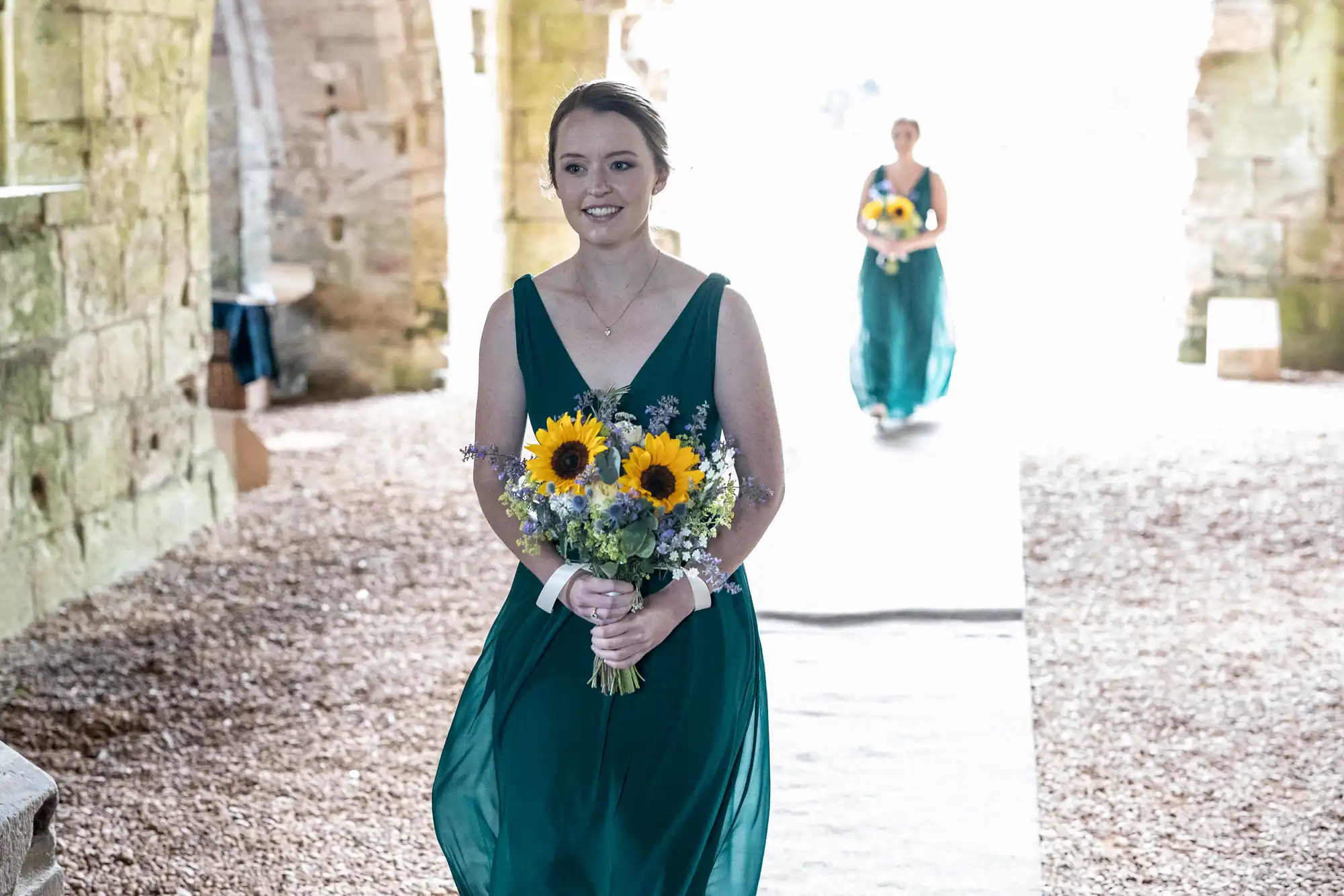 A bridesmaid in a green dress holding a bouquet of sunflowers walks through a stone archway, followed by another bridesmaid in the distance.