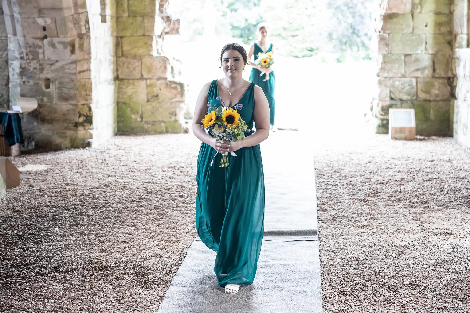 A woman in a teal dress holds a bouquet of sunflowers while walking down an aisle in a stone building, followed by another woman carrying a bouquet.