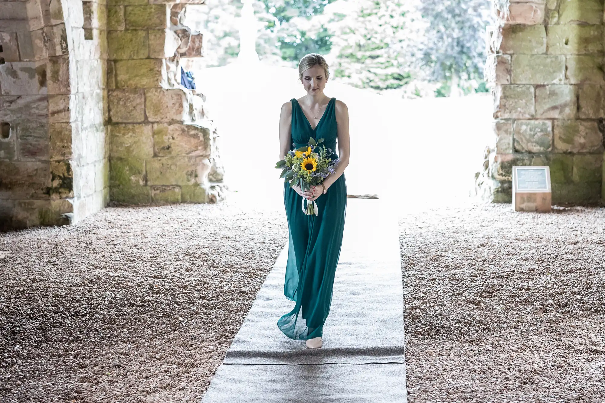 A woman in a green dress walks down an aisle holding a sunflower bouquet, under an archway with stone walls and a bright, natural background.