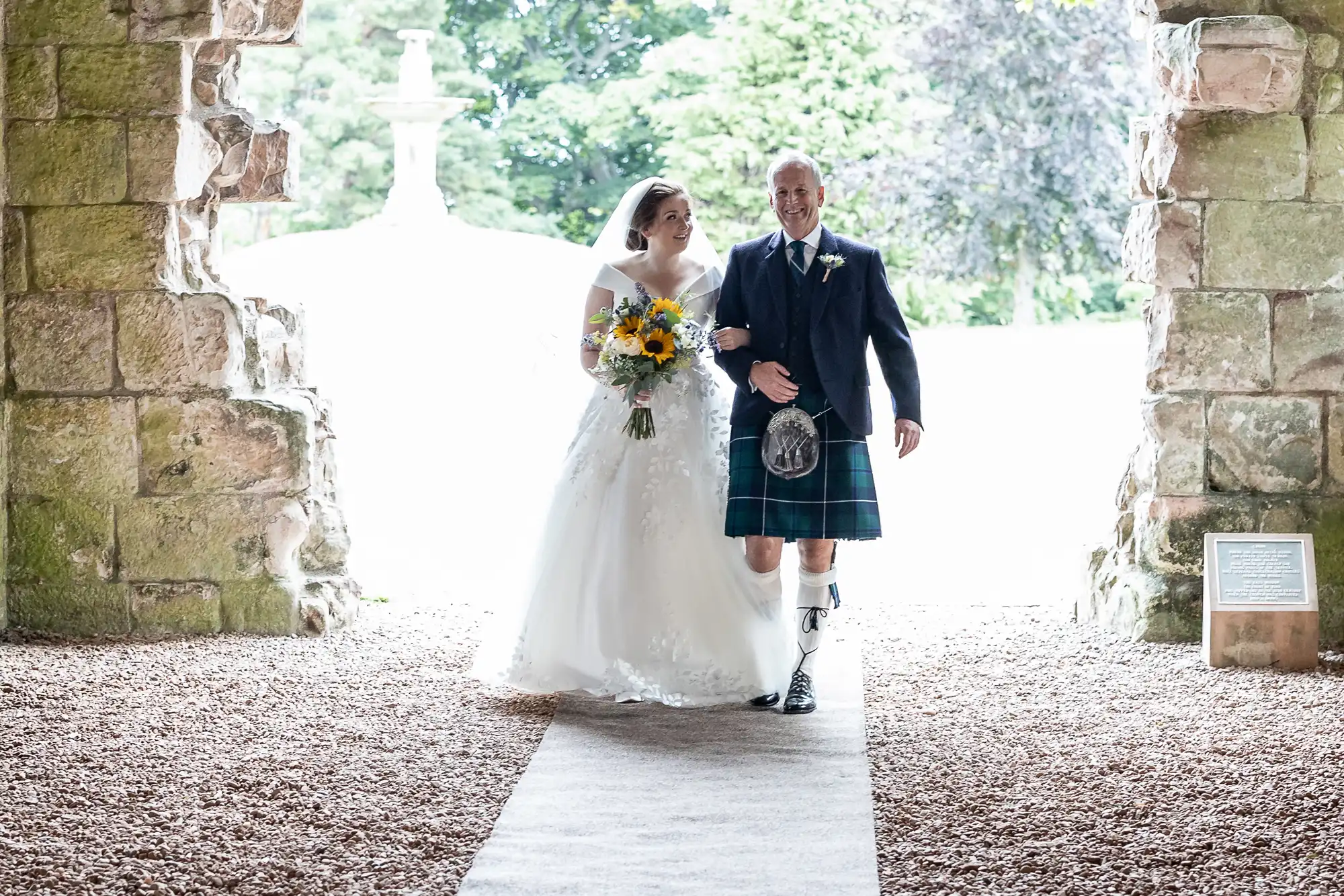 Bride in a white gown holding sunflowers walks arm-in-arm with a man in a kilt, entering through an old stone archway.