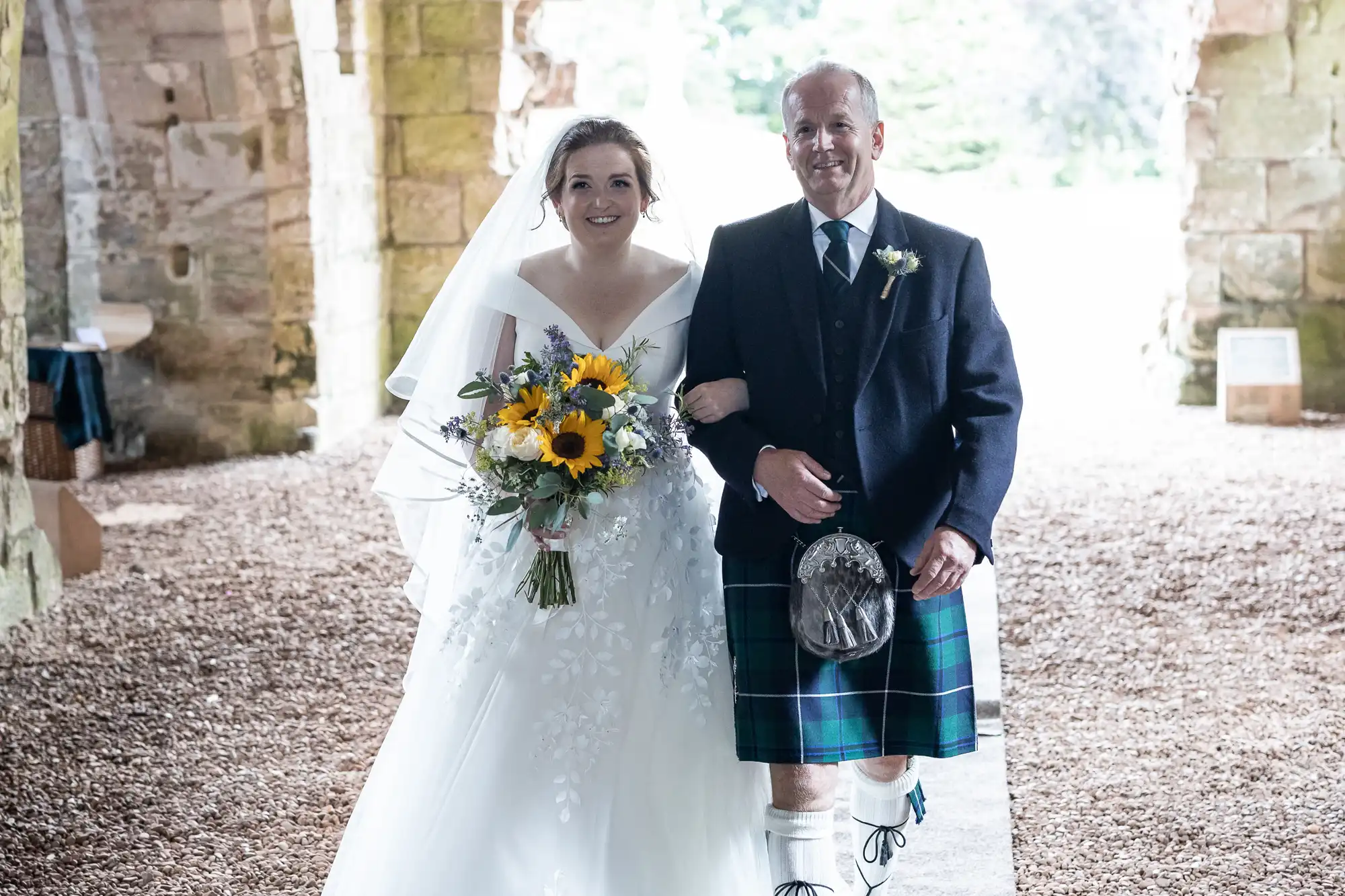 A bride in a white gown and veil holds a bouquet of sunflowers, walking with a man in a kilt and dark jacket inside an old stone building.