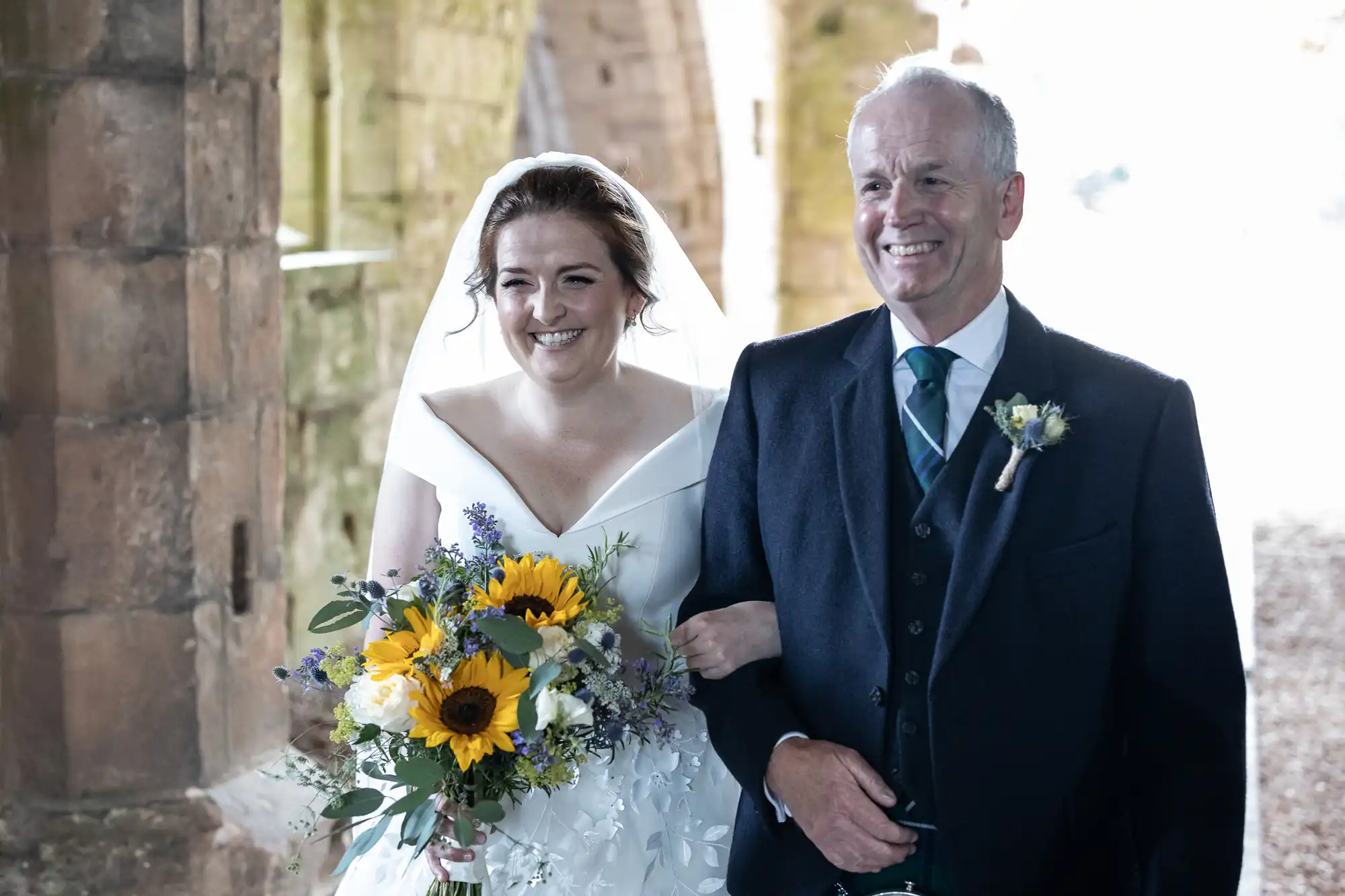 A bride in a white dress holding a bouquet of flowers and a man in a suit are walking together and smiling. They are in an outdoor setting with stone arches behind them.