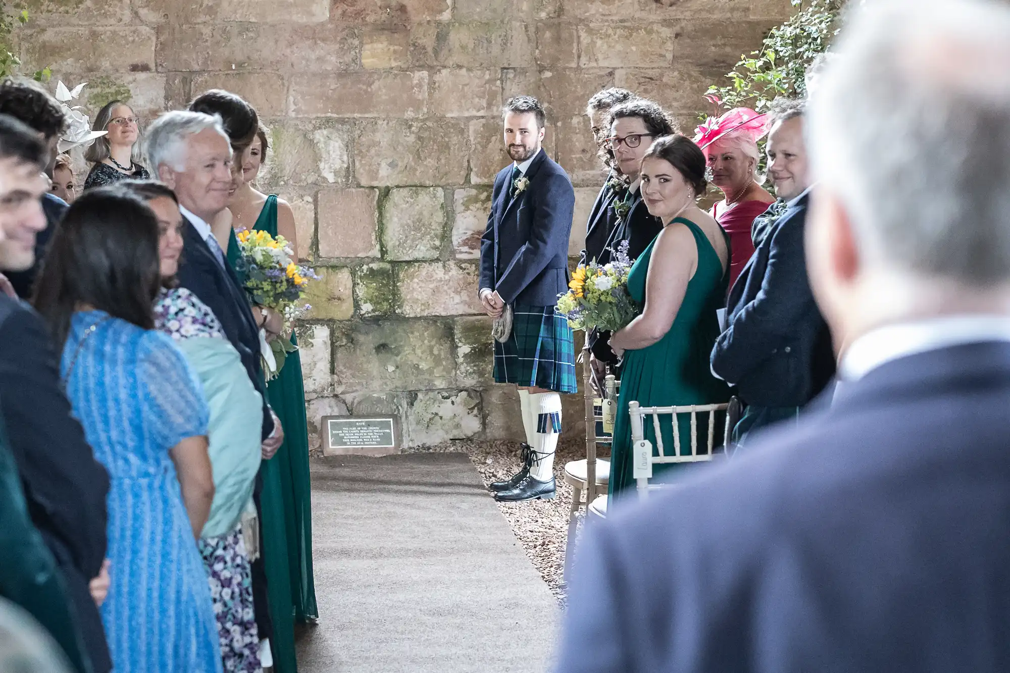 A groom in a kilt stands at the altar, smiling at the bride walking down the aisle. Guests are seated on both sides, turning to look at the bride. The setting is rustic with a stone wall background.