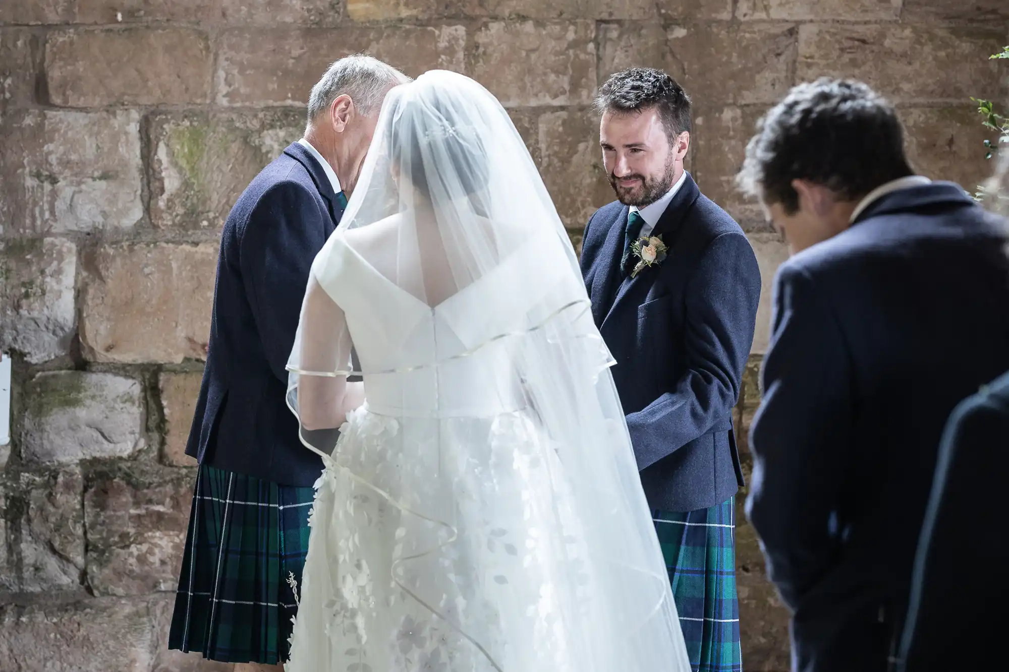 A bride and groom exchange vows, facing each other, with two other men in kilts standing nearby, against a stone wall background.