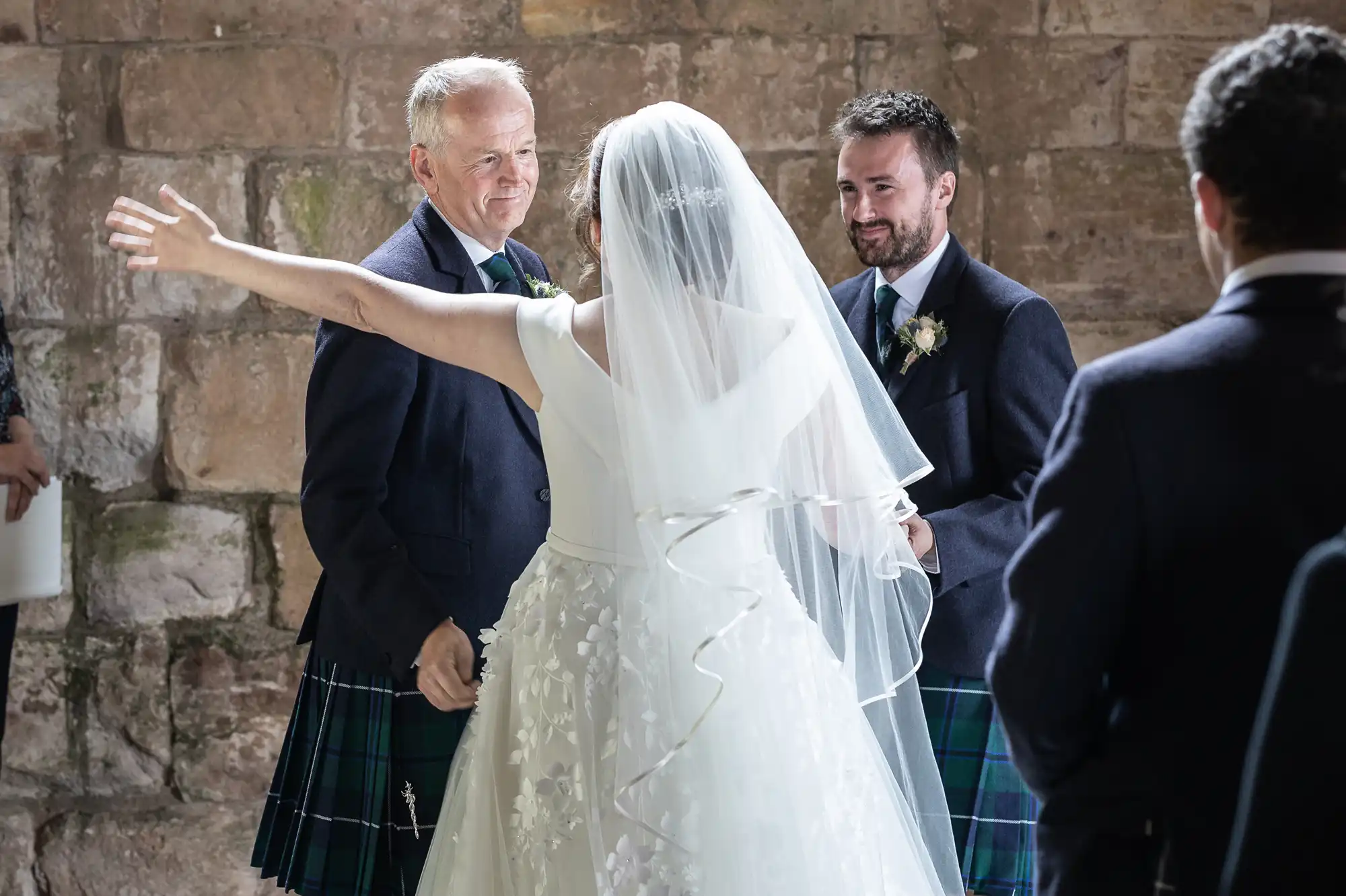 A bride in a white dress with a flowing veil has her arms outstretched toward two men wearing kilts. They are standing in front of a stone wall, and one man smiles while holding her hand.