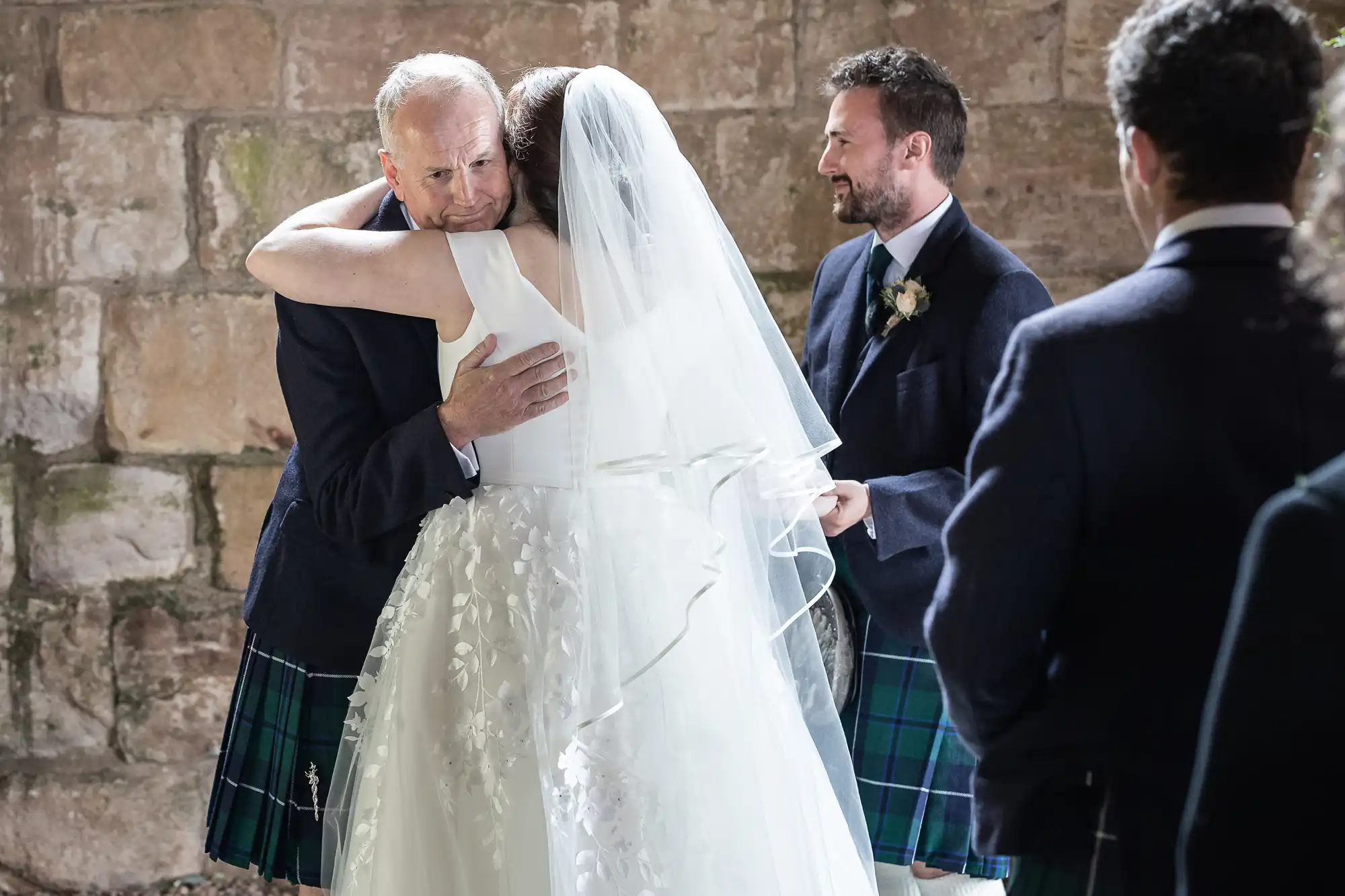 A bride in a white dress and veil hugs an older man in a kilt at a wedding ceremony, with two other men in kilts looking on. They are standing in front of a stone wall.