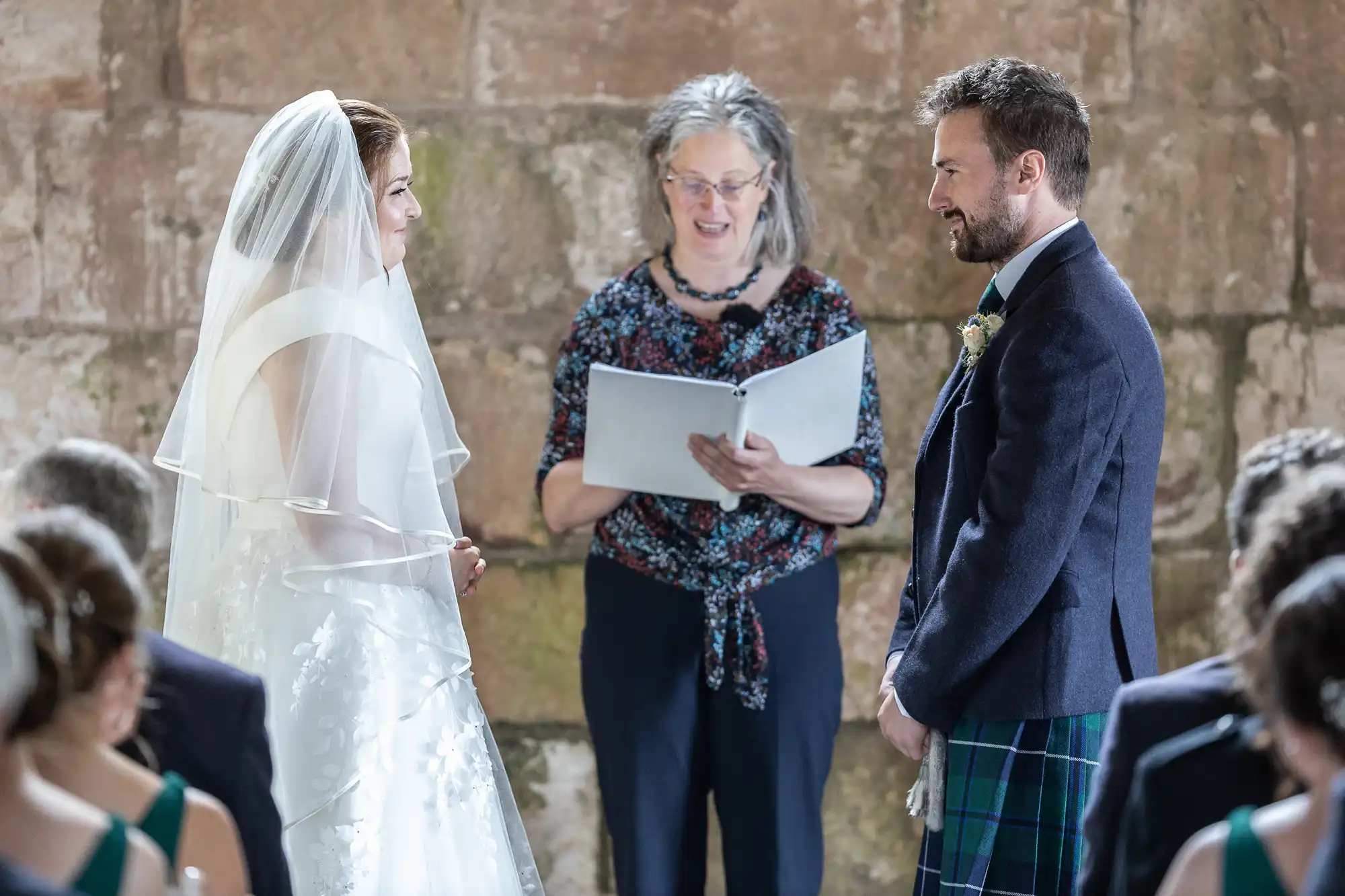 A bride and groom stand facing each other during their wedding ceremony while an officiant reads from a book. Guests are seated in the foreground.