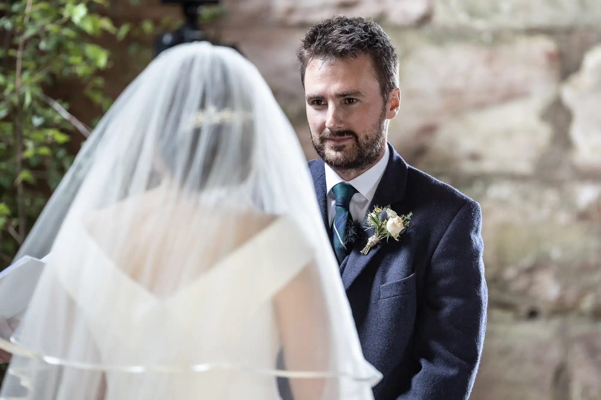 A groom in a suit and tie looks at a bride in a wedding dress with a veil, both standing indoors near a stone wall and greenery.