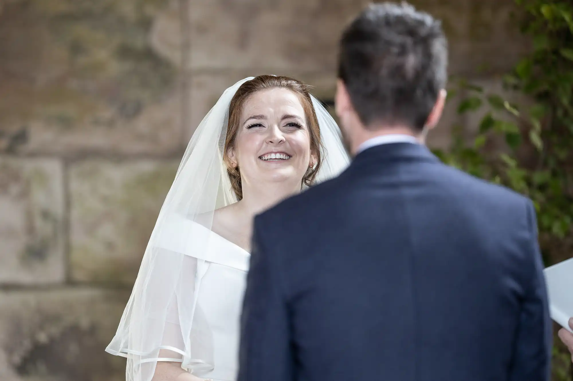 A bride and groom stand facing each other during their wedding ceremony; the bride is smiling, and the groom's back is facing the camera.