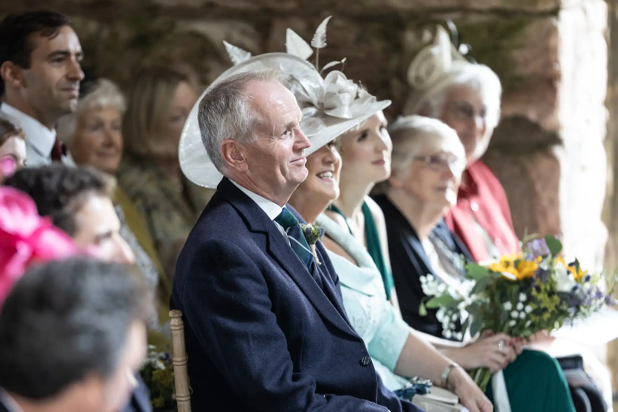 A group of people, dressed in formal attire and hats, sit closely together, likely attending a wedding or formal event, with one man in the foreground wearing a dark suit and tie.