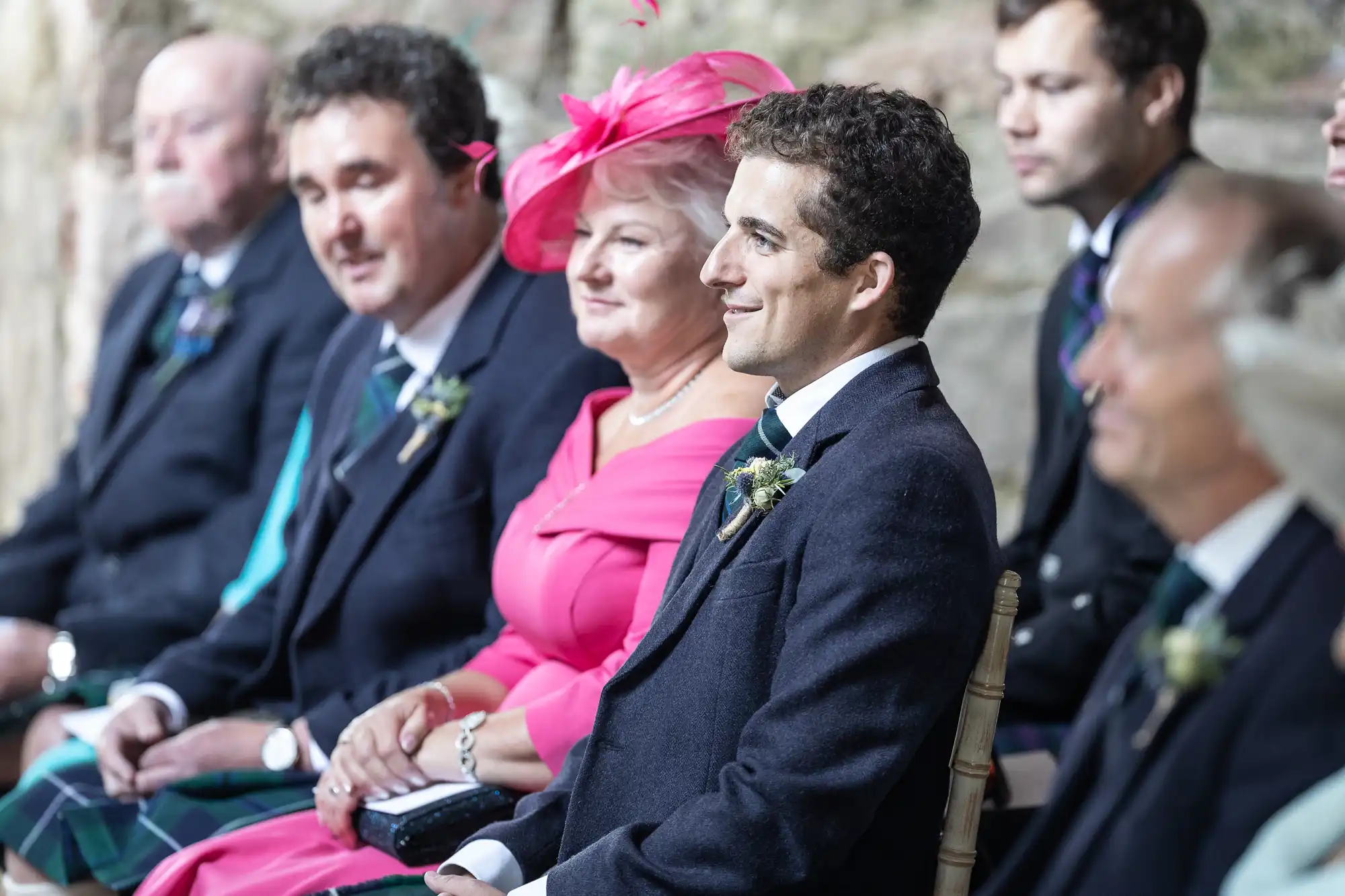 A group of people, dressed formally, are seated in a row. One woman is wearing a bright pink outfit and hat, while the others are in dark suits with boutonnieres.