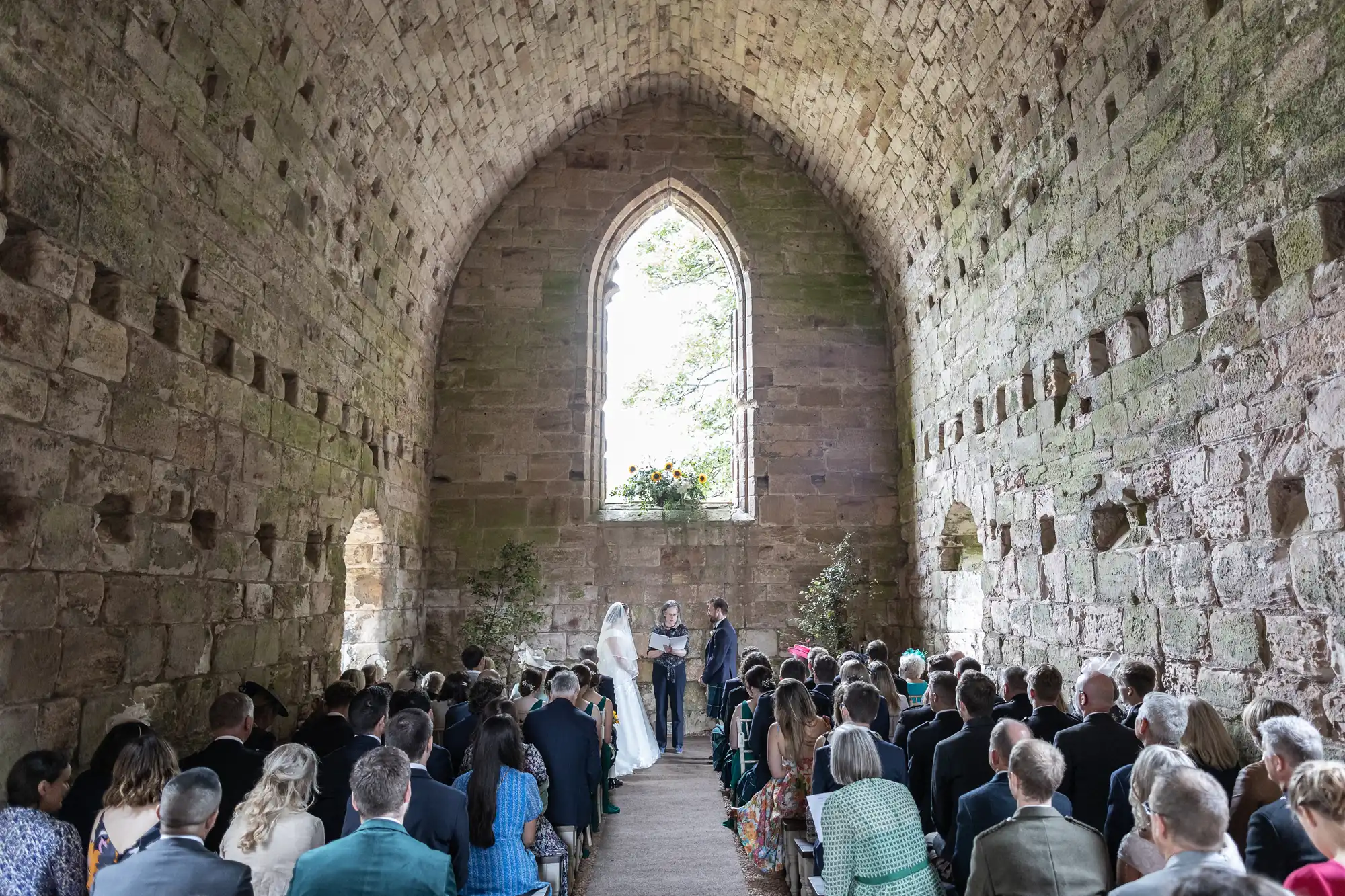 A wedding ceremony taking place inside an old stone chapel, with guests seated on both sides, and a couple standing at the altar under a large arched window.